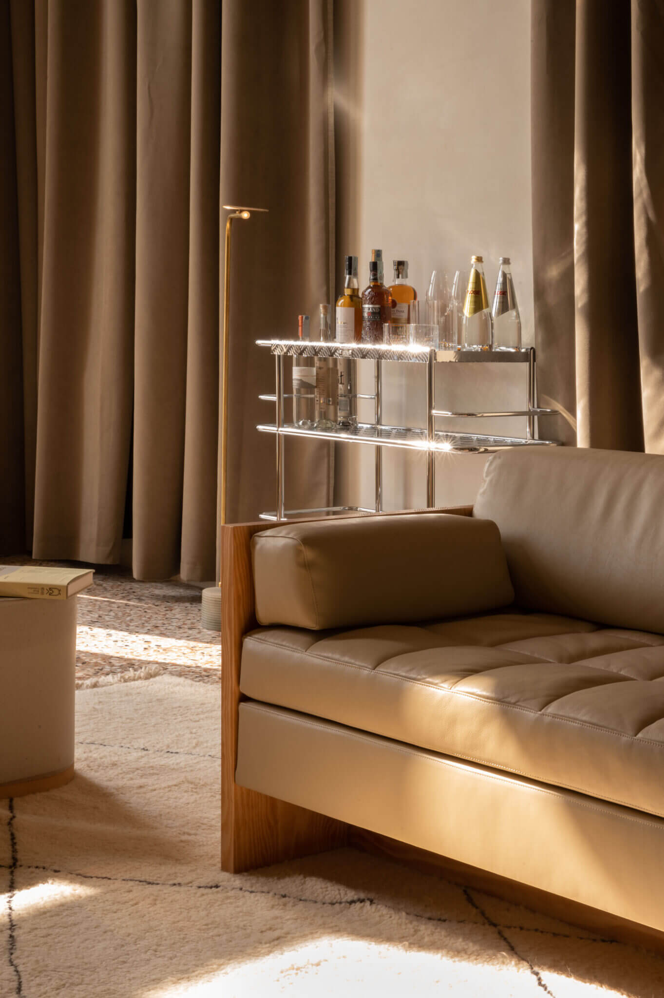 The Venice Venice Hotel, Europe — Minimalist bar cart with illuminated shelves displaying spirits and glassware beside a leather sofa under warm natural light filtering through floor-to-ceiling curtains.