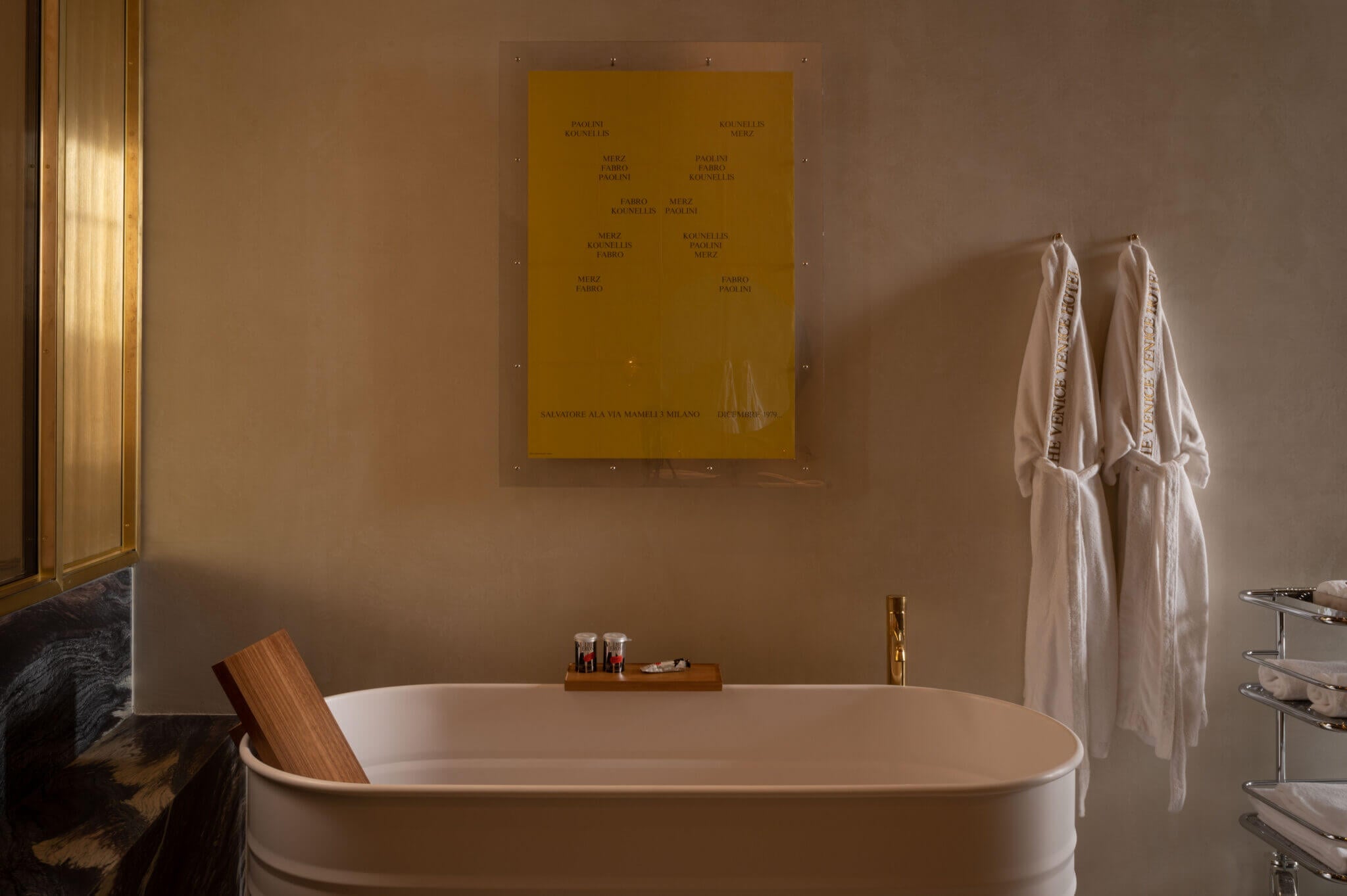 The Venice Venice Hotel, Europe — minimalist spa bathroom with oval soaking tub, warm taupe walls, golden artwork, wooden headrest, cream linens, brass fixtures, and soft ambient lighting.