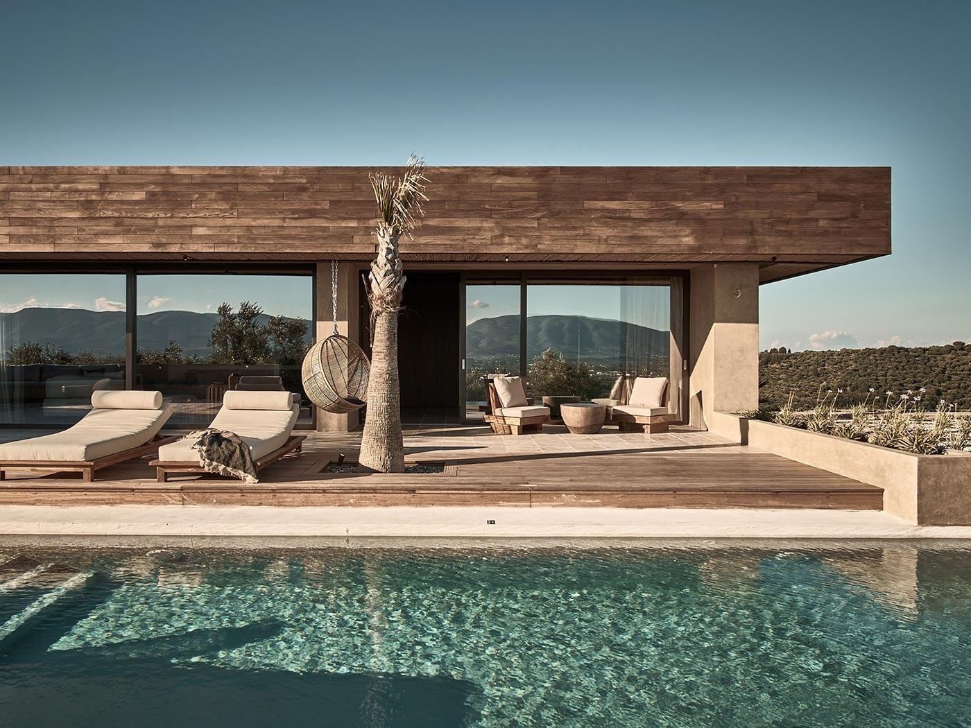 Olea All Suite Hotel, Europe — Modern poolside pavilion with wooden deck, lounge chairs, palm tree, and mountain views under clear sky.