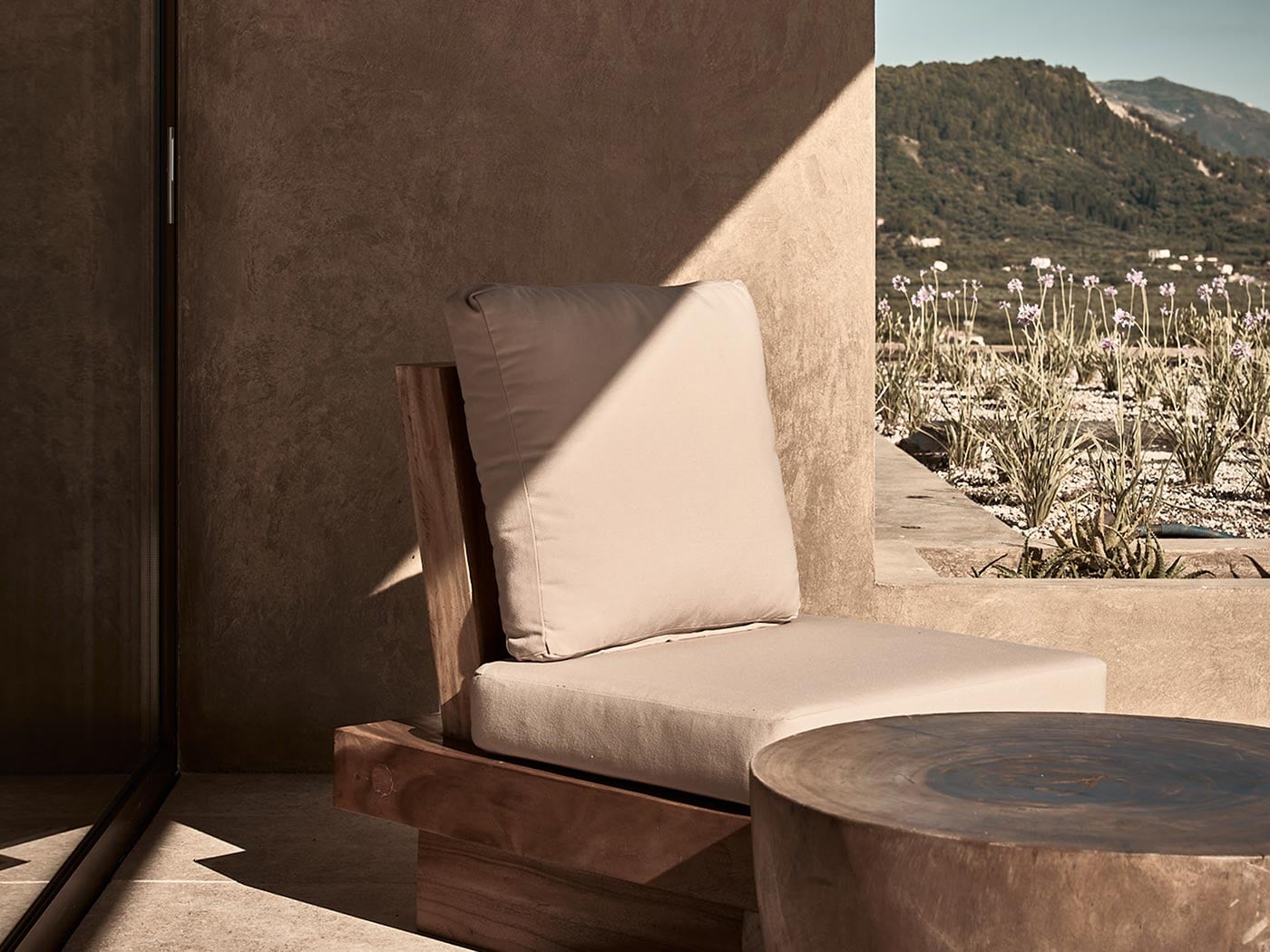 Olea All Suite Hotel, Europe — Minimalist wooden lounge chair with cream cushions and pillow on stone terrace, morning light casting shadows, wildflower field and forested mountains beyond.