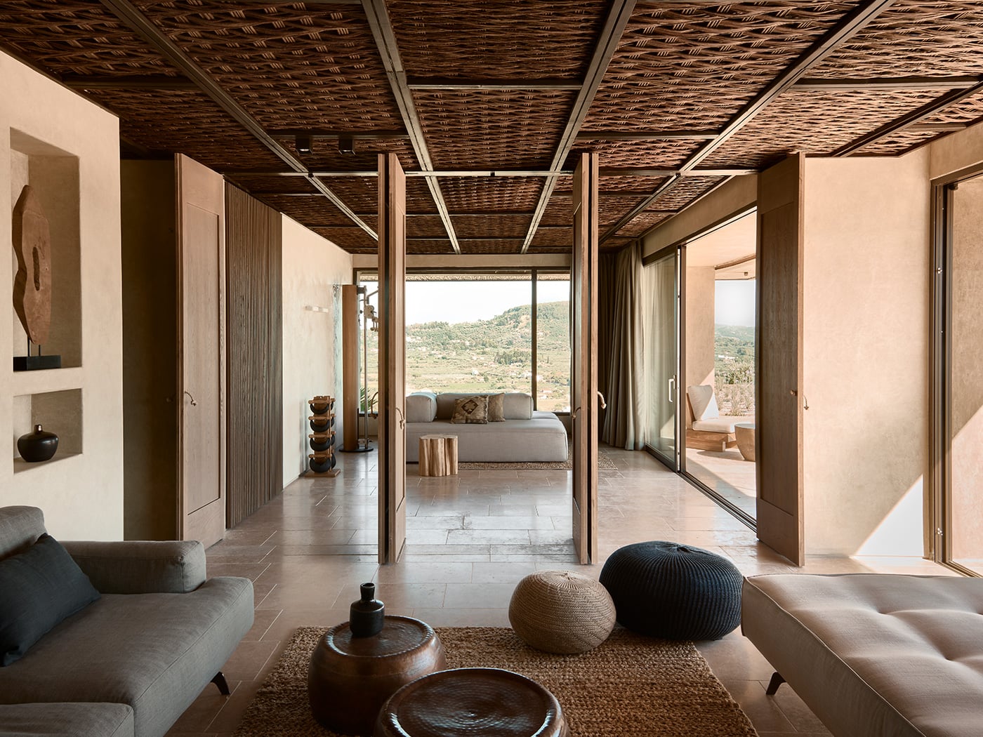 Olea All Suite Hotel, Europe — Minimalist suite with woven ceiling, stone floors, earth-toned walls, panoramic landscape views through floor-to-ceiling glass doors, natural light, neutral furnishings.