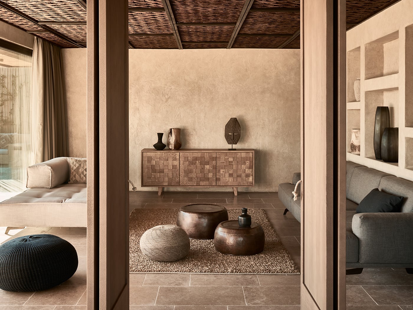 Olea All Suite Hotel, Europe — Minimalist spa lounge with woven ceiling, textured walls, low seating poufs, wooden sideboard, and earthy neutral palette in warm natural light.