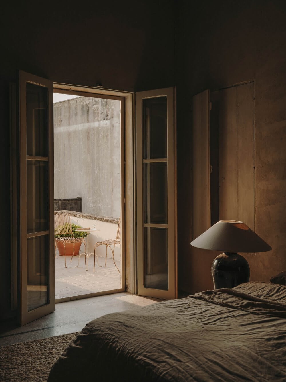 Casa Soleto, Europe — Terracotta bedroom with open French doors to sunlit courtyard, minimalist table lamp, linen bedding, and weathered stone walls.