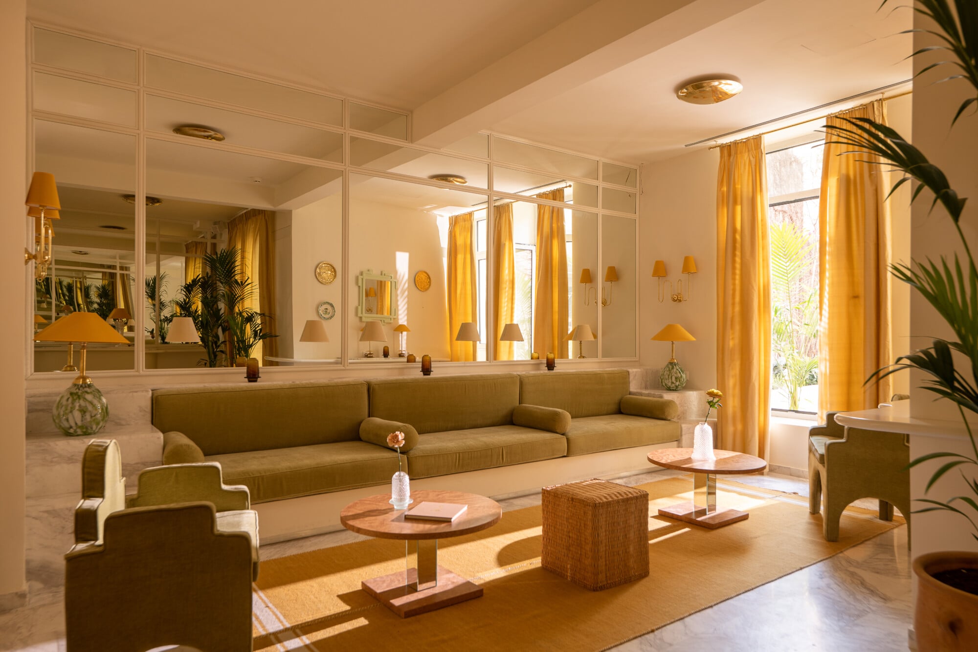 Apollo Palm, Europe — Sunlit modernist living room with olive-green sectional sofa, mirrored walls, golden curtains, wooden coffee table, and potted palms.
