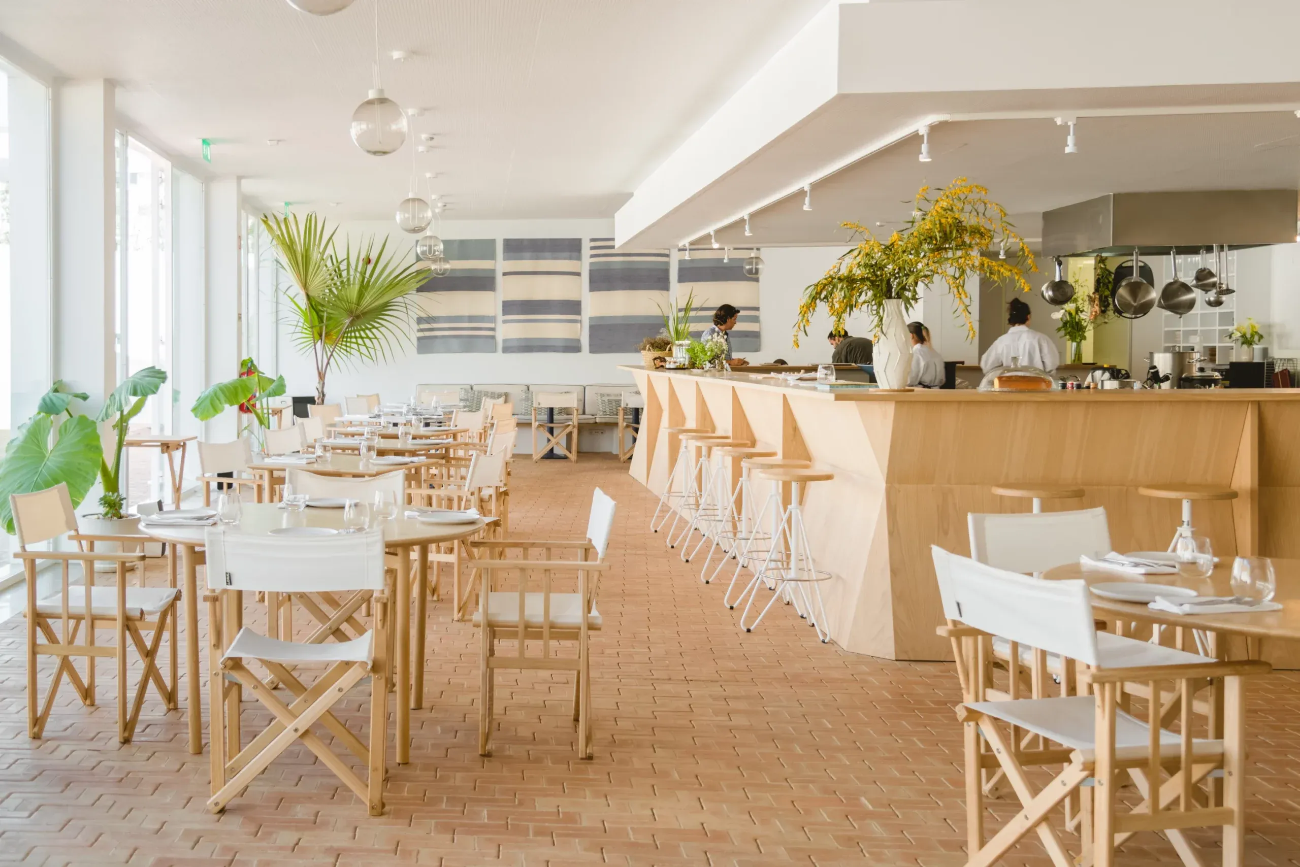 Casa Mãe, Europe — Bright dining room with natural wood furniture, striped backdrop, pendant lighting, lush plants, and open kitchen with staff.