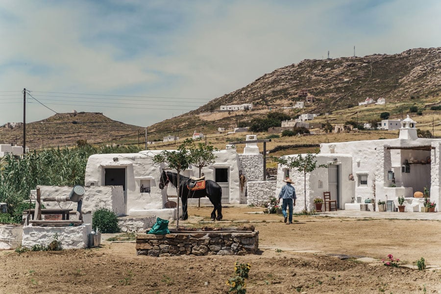 Rizes Folklore Farmstead, Europe — Whitewashed stone buildings with terracotta roofing nestled on arid hillside, horse and figure in dusty courtyard, Mediterranean vernacular architecture.