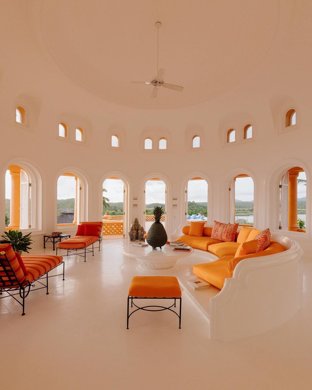 Cuixmala, Mexico — Curved cream interior with arched windows, orange upholstered seating, black iron furniture, and river valley views through colonnade openings.