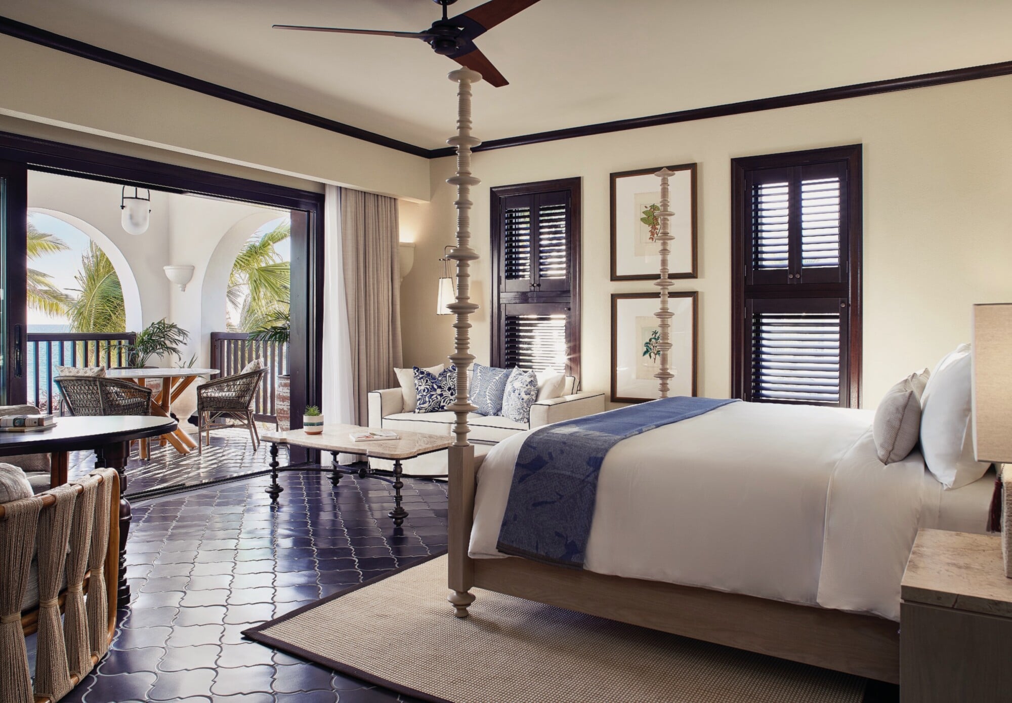 Cap Juluca, North America — Coastal resort bedroom with four-poster bed, dark tile floors, arched balcony doors, cream walls, shuttered windows, and ocean views through colonnade.