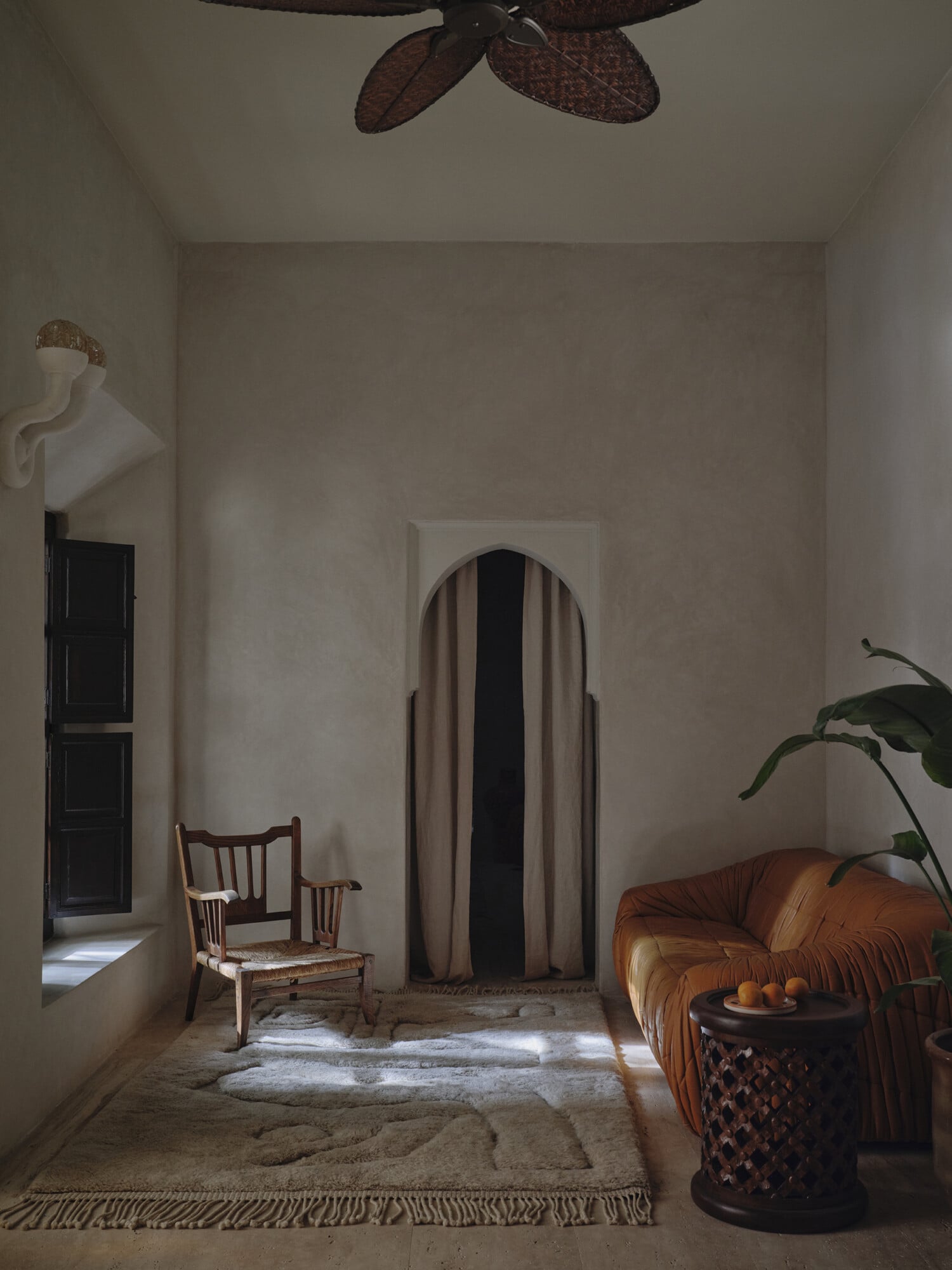 Rosemary, Africa — Minimalist parlor with arched doorway, cognac leather sofa, woven chair, ceramic side table, potted plant, and rattan ceiling fan casting shadows.