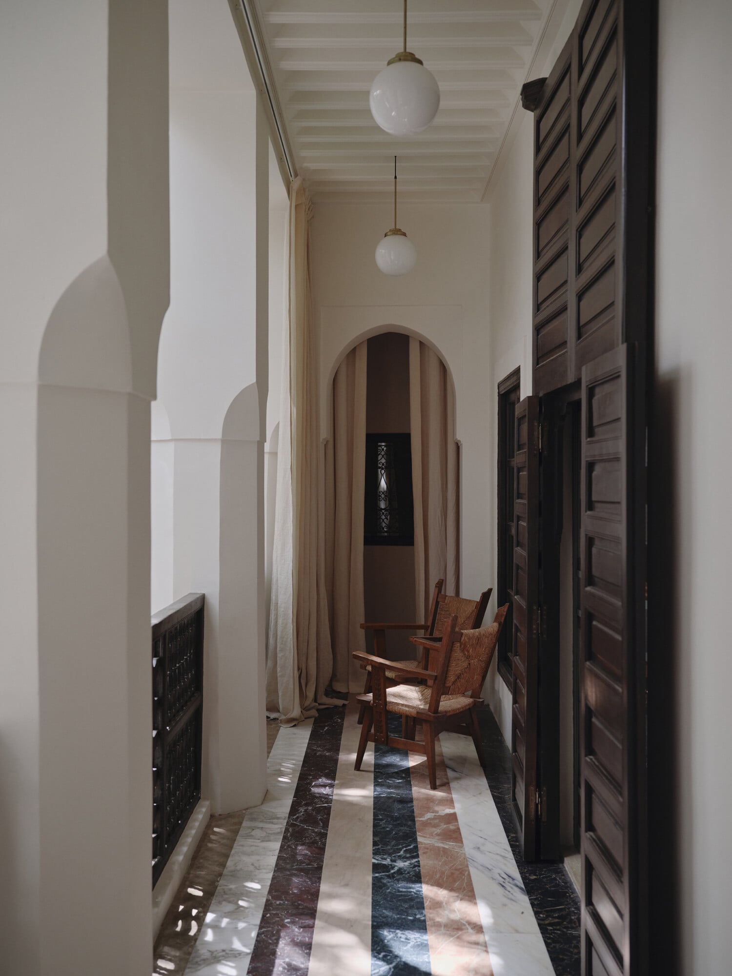 Rosemary, Africa — Narrow corridor with striped marble floor, arched doorway, pendant lights, wooden chairs, dark carved doors, minimalist aesthetic.