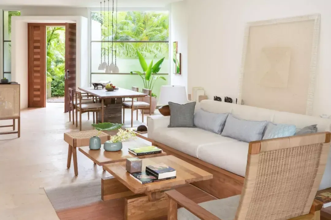 Rosewood Mayakoba, Mexico — Open-plan living room with teak furnishings, rattan details, cream upholstery, and floor-to-ceiling windows overlooking tropical garden views.