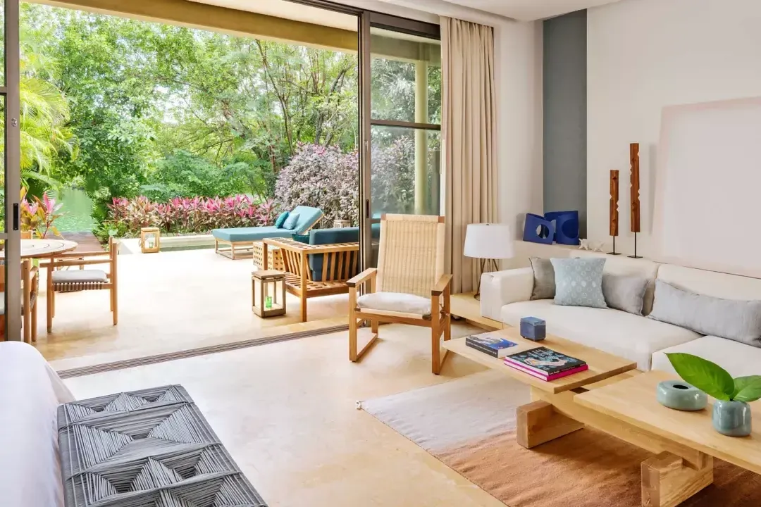 Rosewood Mayakoba, Mexico — Modern living room with wooden furniture, neutral palette, opening to lush garden terrace with flowering plants and lounge seating.