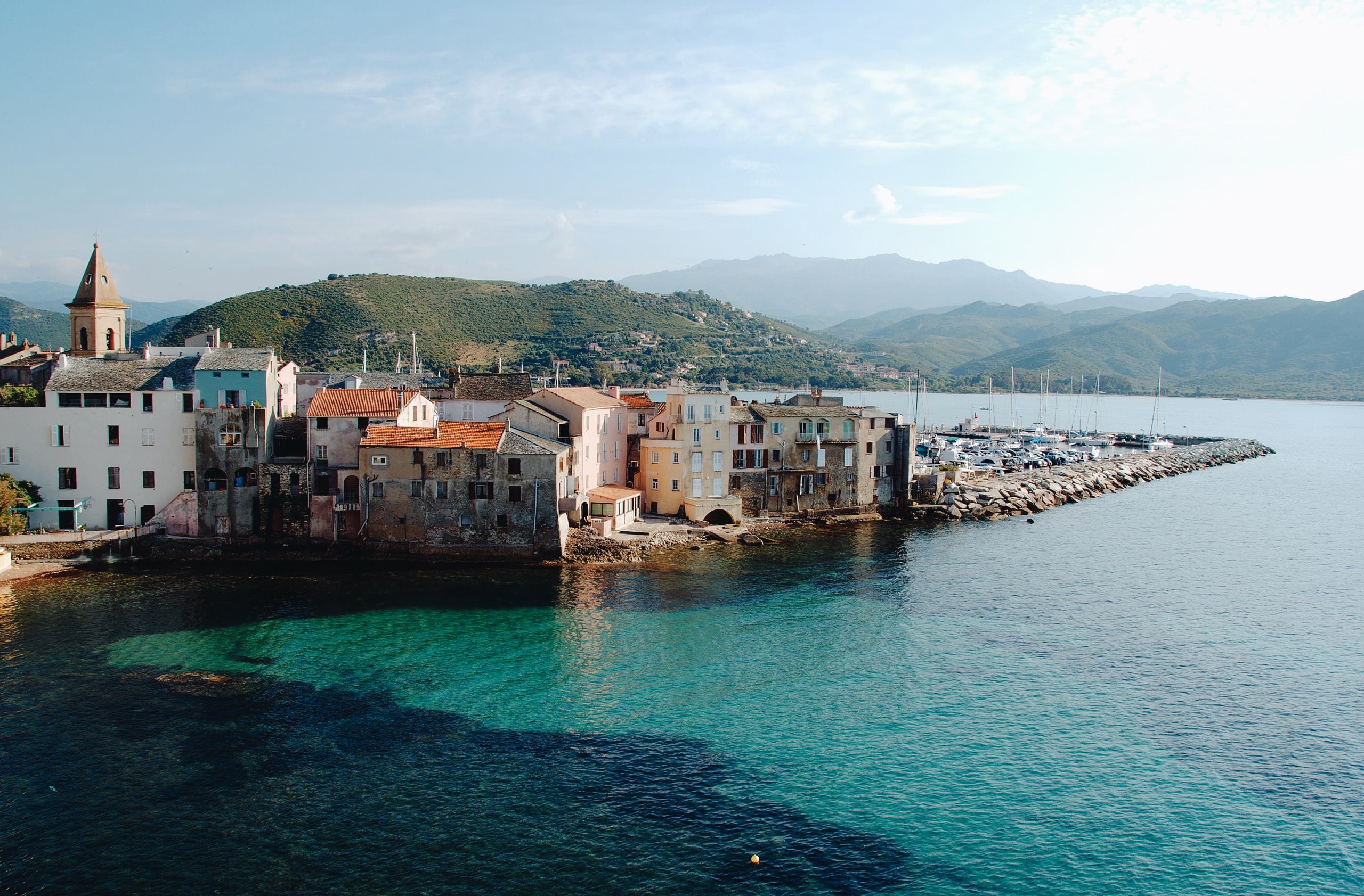 Saint-Florent, Corsica, France — Pastel waterfront village with church bell tower, colorful buildings along turquoise Mediterranean cove, forested mountains, moored sailboats.