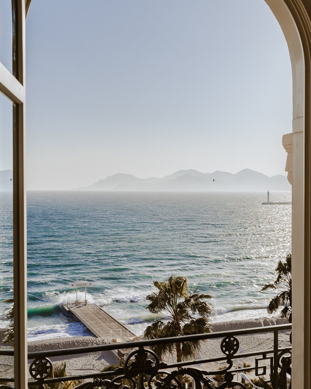 Carlton Cannes, Cannes, France — Beachfront view through arched doors with wrought-iron balcony, Mediterranean Sea, distant mountains, palm fronds, morning light.