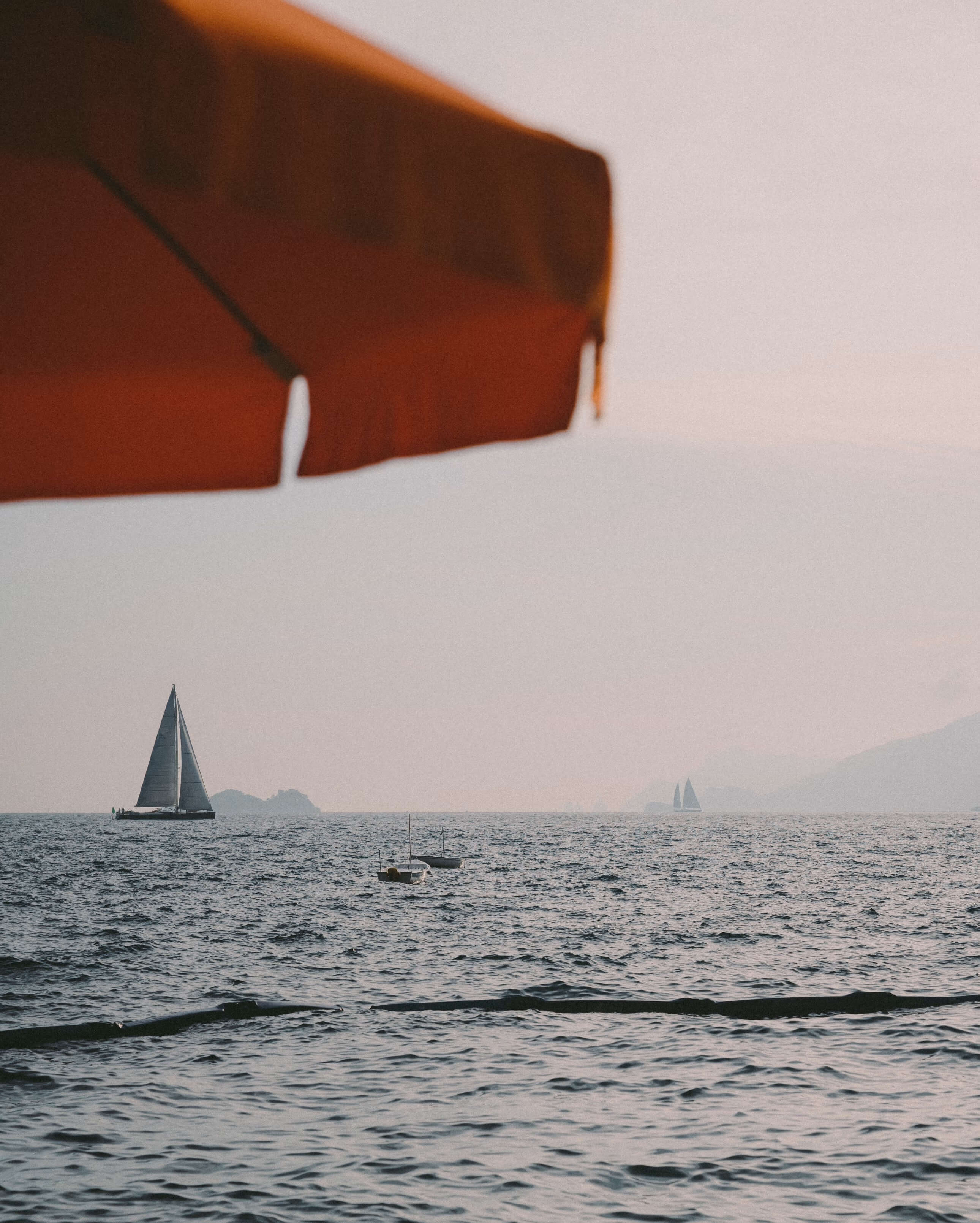 Splendido Mare, Europe — Rust-colored beach umbrella framing sailboats on misty waters at dusk, coastal luxury aesthetic.
