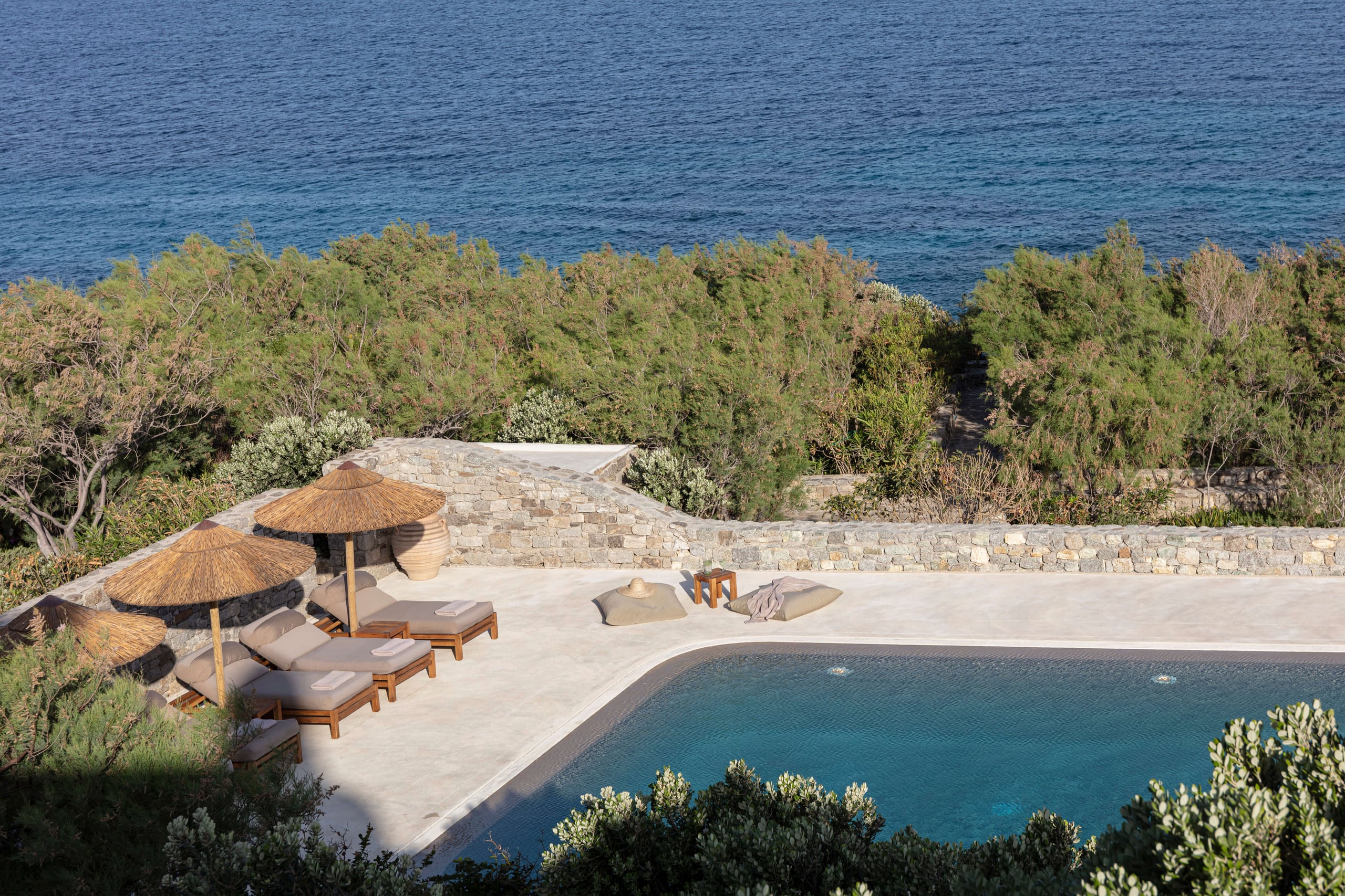 Villa Tama, Europe — Coastal terrace with thatched umbrellas, loungers, stone fireplace, and infinity pool overlooking Mediterranean sea and pine forest.