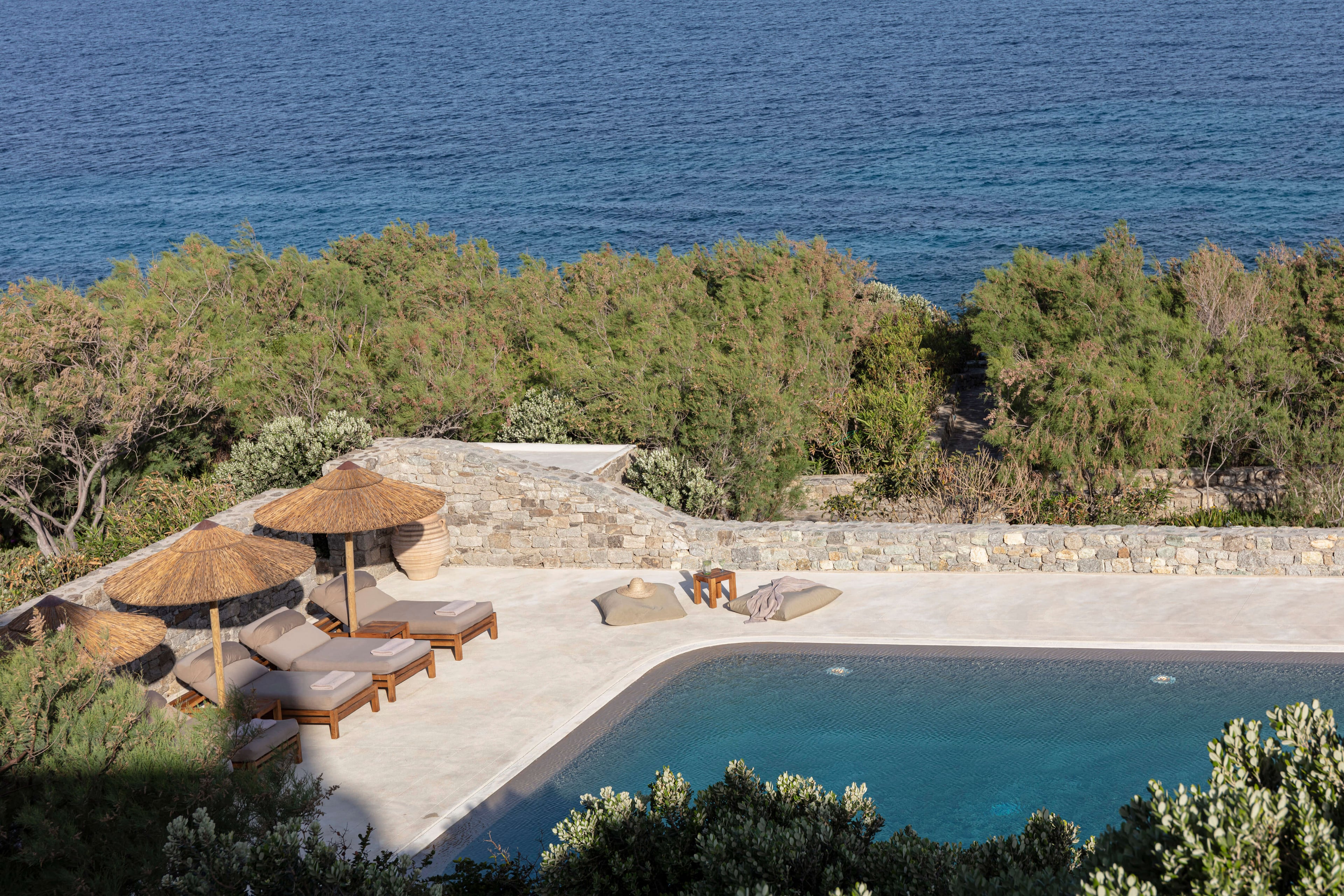 Villa Tama, Europe — Coastal terrace with thatched umbrellas, loungers, stone fireplace, and infinity pool overlooking Mediterranean sea and pine forest.