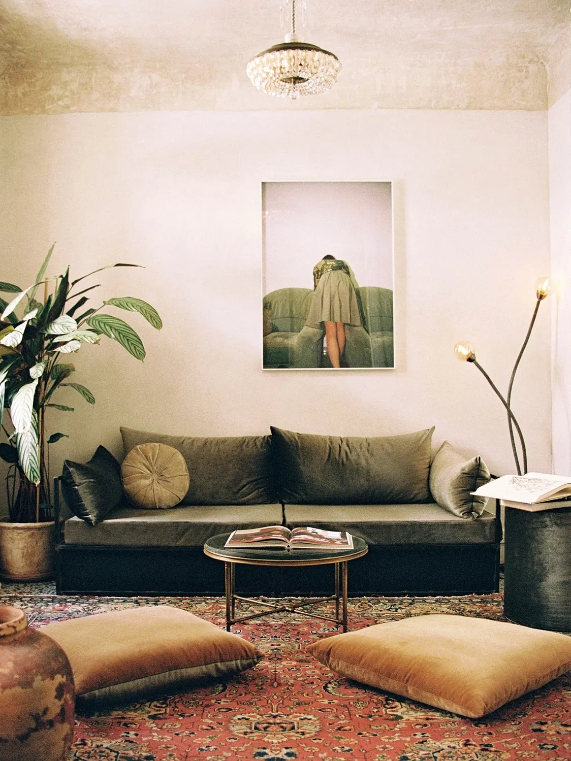 Shila, Europe — Minimalist living room with olive linen sofa, brass floor lamp, patterned rug, potted plants, and framed portrait above round side table.