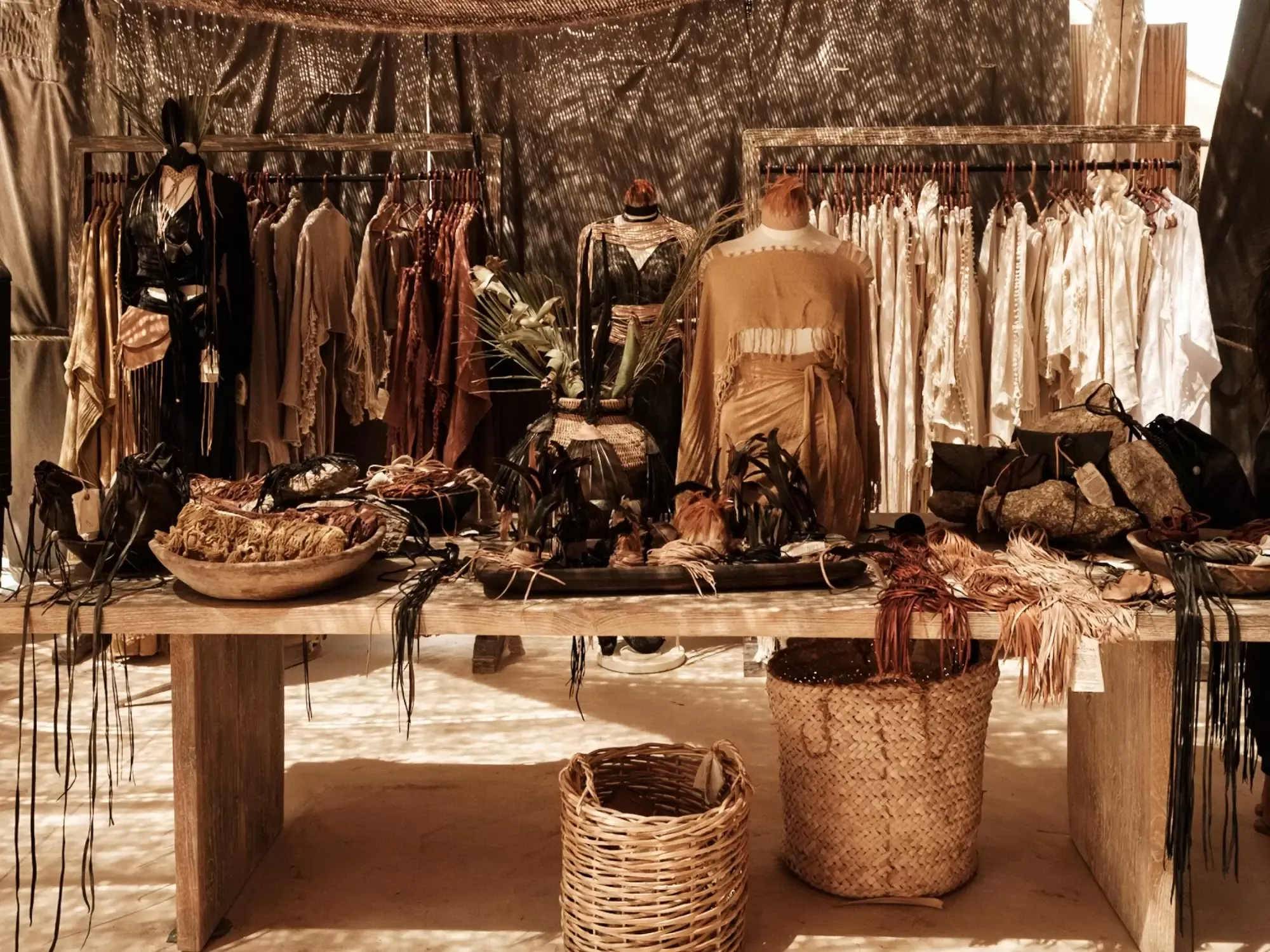 Caravana, Mexico — Earthy textile display with natural linen clothing, woven baskets, dried botanicals, and handcrafted accessories arranged on wooden tables under mesh canopy.