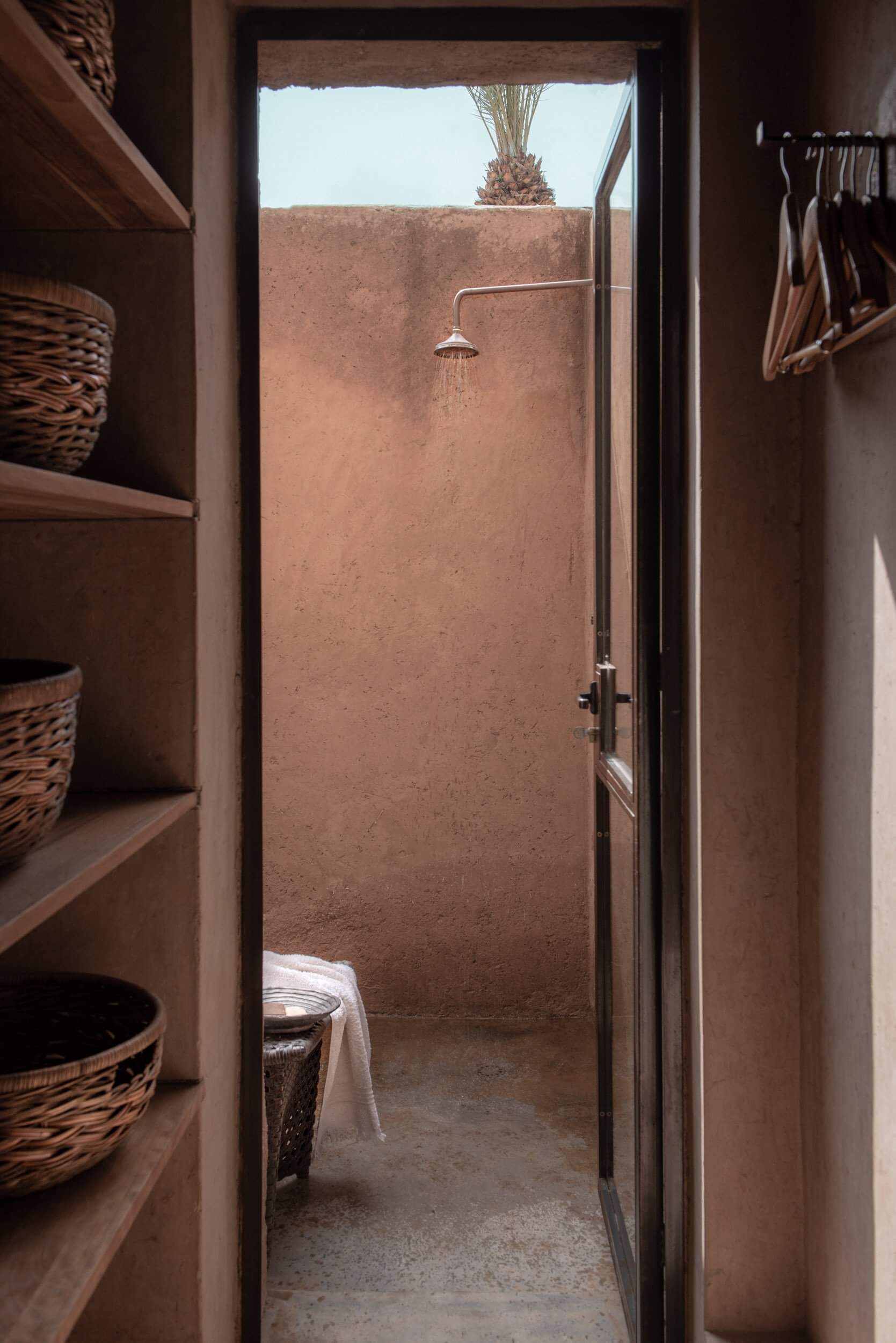 Villa D, Africa — Open-air shower with clay walls, palm tree views, woven storage baskets, minimalist design and natural light.