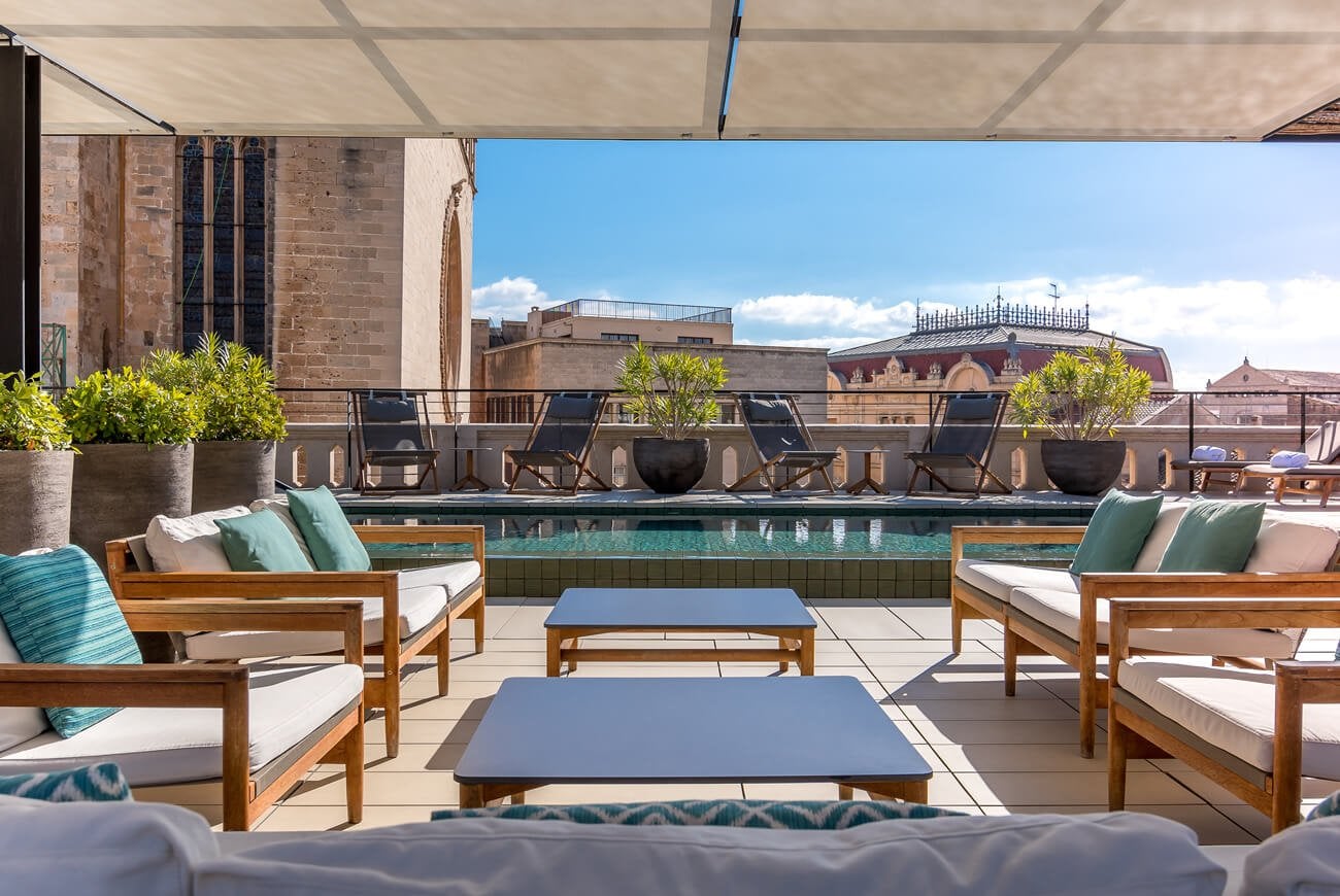 Singular Rooftop, Balearic Islands, Spain — Wooden lounge seating with turquoise pool overlooking historic brick architecture under retractable canopy.