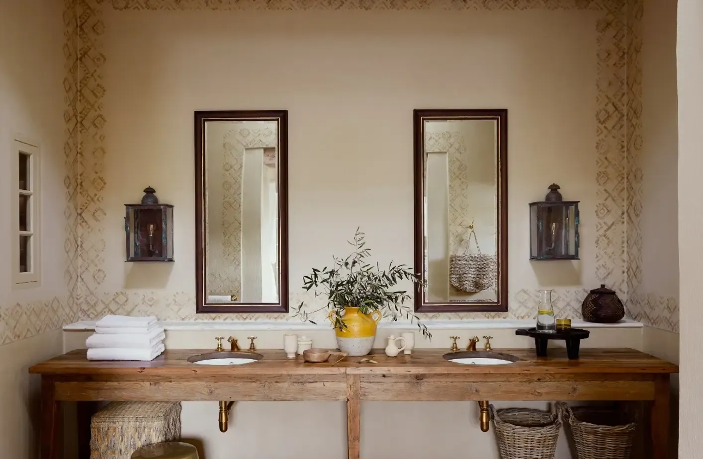 Sterrekopje, Africa — Double vanity with reclaimed wood counter, brass fixtures, patterned wallpaper, framed mirrors, and botanical arrangement in yellow pitcher.