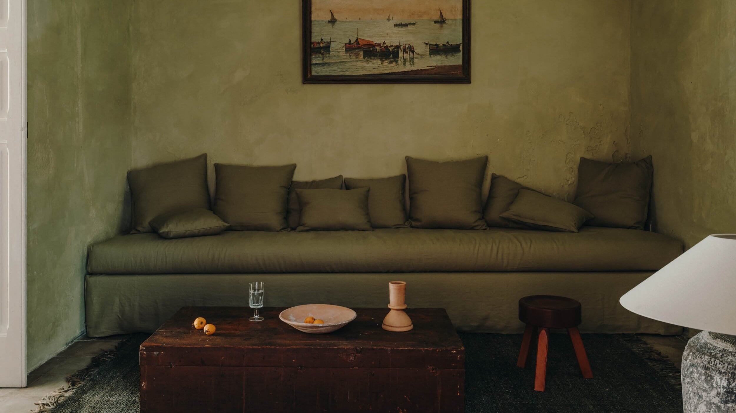 Casa Soleto, Europe — Olive linen sofa with throw pillows against textured sage wall, dark wood table, ceramic vessels, and framed maritime painting above.