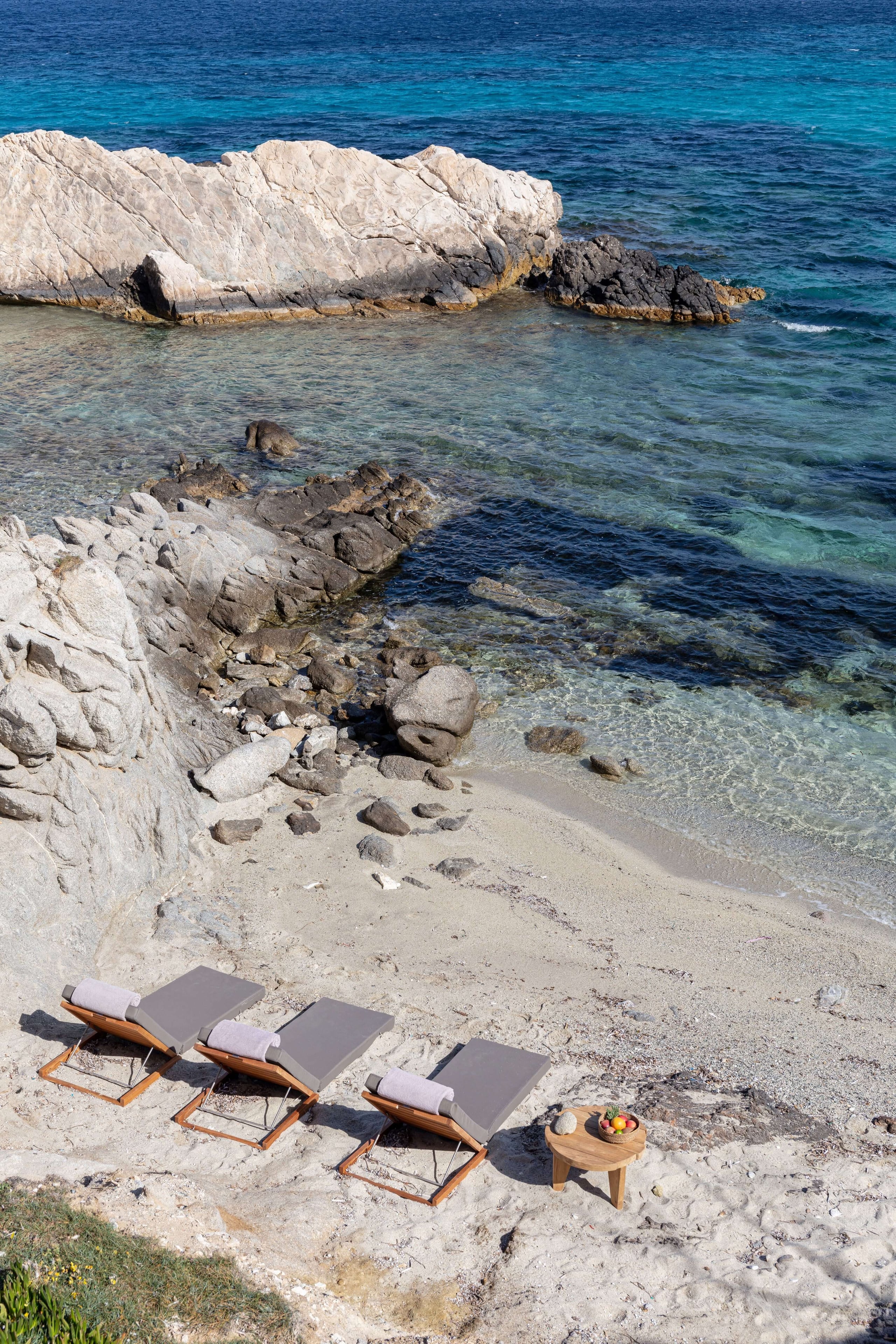 Villa Tama, Europe — Three teak loungers with gray cushions on white sand beach beside turquoise Mediterranean waters and pale limestone cliffs.