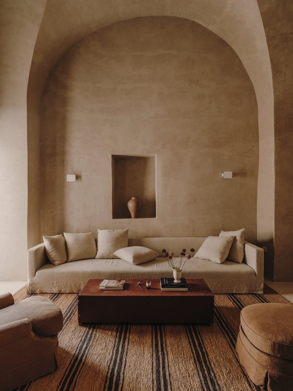 Casa Soleto, Europe — Arched plaster alcove with curved sofa, terracotta vessels, striped natural fiber rug, and dark wood coffee table in minimalist Mediterranean interior.