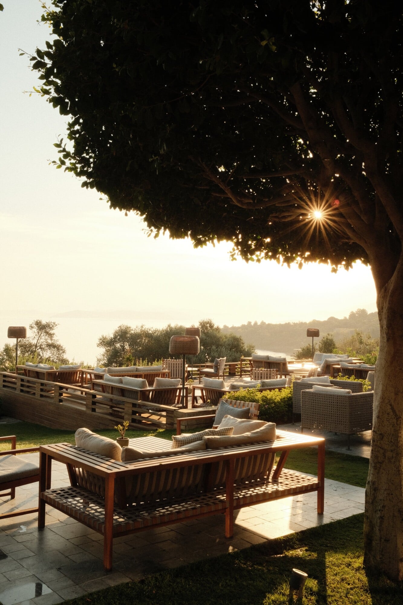 Sofra, Asia — Waterfront terrace at sunset with wooden lounge furniture, mature shade tree, and distant lake view through manicured gardens.