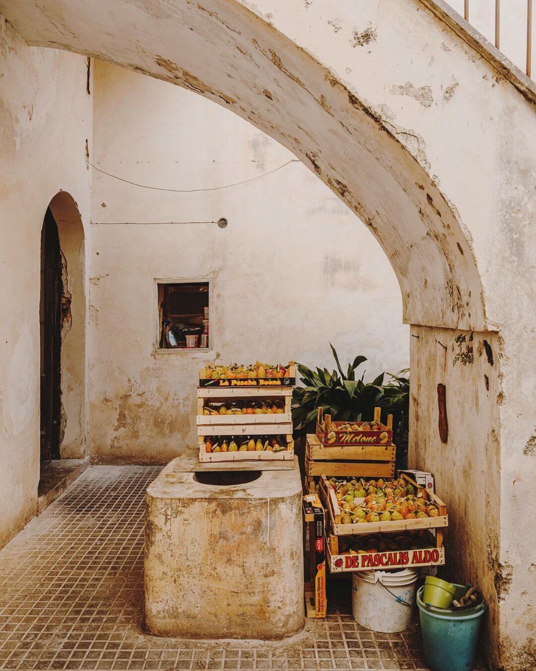 Soleto, Italy — Weathered archway frames stacked produce crates and potted plants in sun-drenched Mediterranean courtyard with terracotta tile floor.