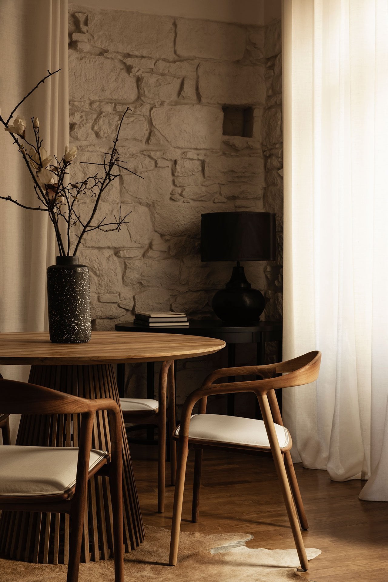 Casa Somos Suenos, Europe — Mid-century wooden dining table and chairs beside brick alcove with black lamp, dried botanicals, and natural light filtering through white curtains.