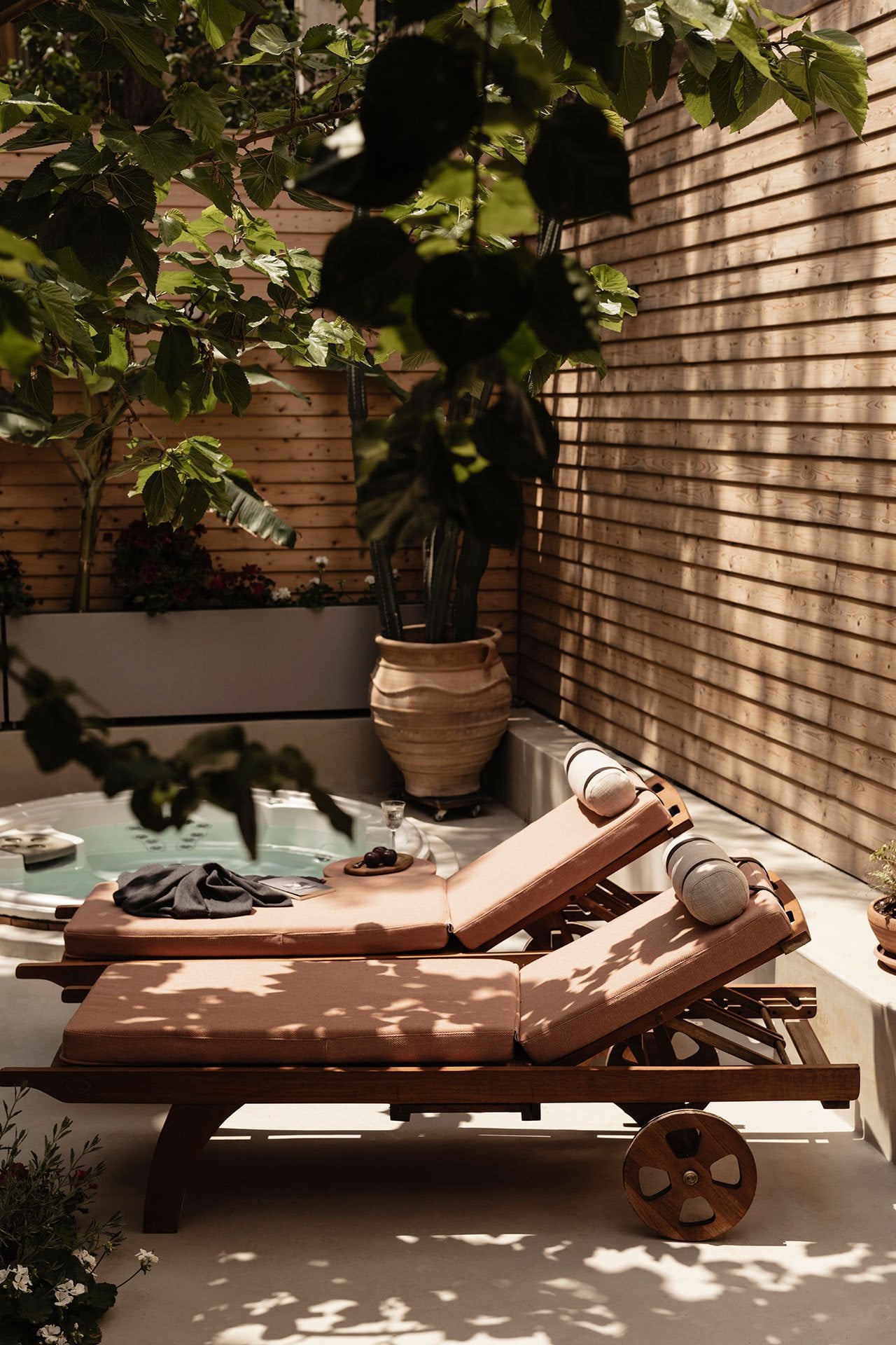 Casa Somos Suenos, Europe — Wooden lounge chairs with cognac cushions on terrace, potted plants, striped brick wall, dappled sunlight through foliage.