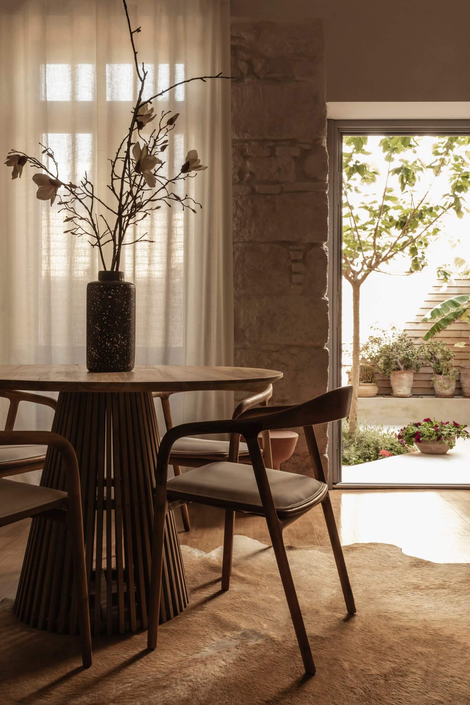 Casa Somos Suenos, Europe — Round wooden dining table with pleated base, curved chairs, dried branches in speckled vase, sheer curtains, terracotta courtyard beyond.