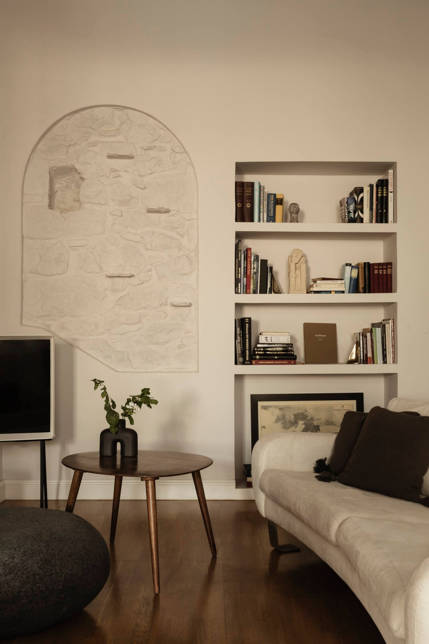 Casa Somos Sueños, Europe — Minimalist bedroom with arched plaster relief artwork, floating shelves, mid-century table, cream sofa, and warm ambient lighting.