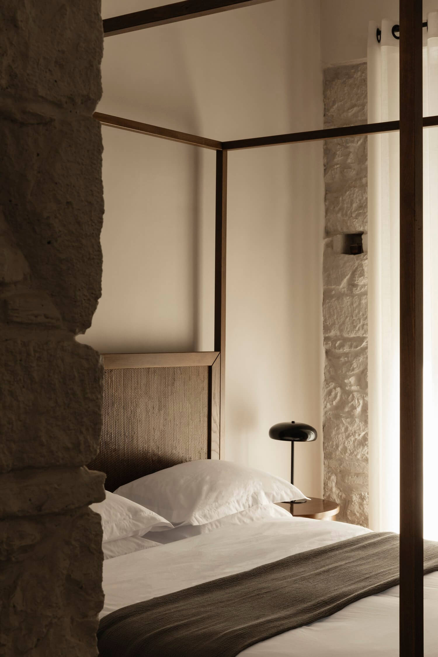 Casa Somos Suenos, Europe — Minimalist canopy bed with dark steel frame, stone walls, white linens, and sculptural bedside lighting in serene bedroom.