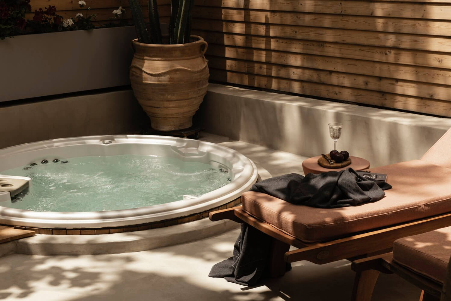 Casa Somos Suenos, Europe — Sunlit spa terrace with round jetted tub, cognac lounger, dark towels, ceramic vessel, and wooden fence casting geometric shadows.