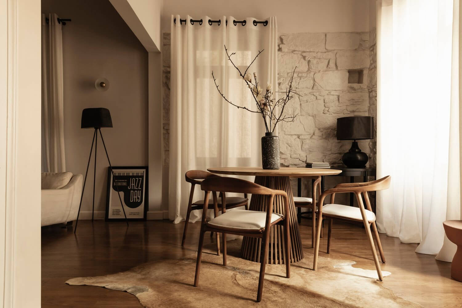 Casa Somos Suenos, Europe — Mid-century dining table with wooden chairs, concrete vase with branches, black tripod lamp, stone accent wall, cream curtains, warm natural light.