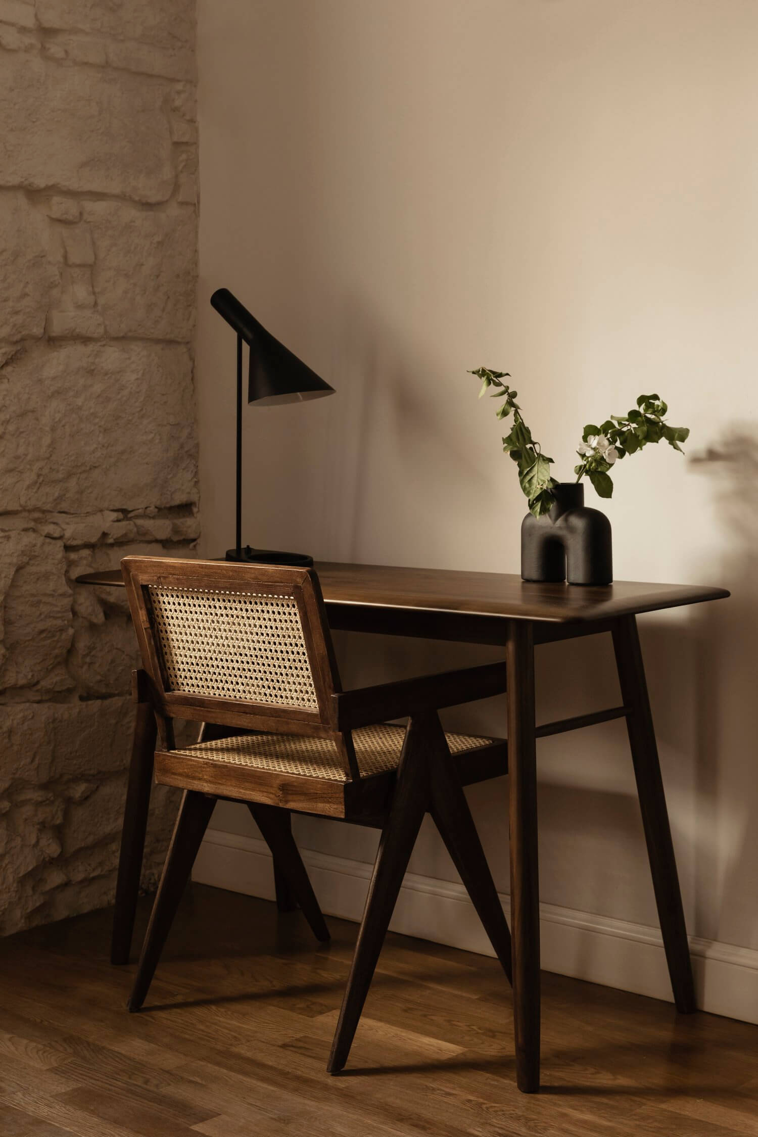 Casa Somos Suenos, Europe — Mid-century desk with rattan chair, black task lamp, potted plant, textured stone wall, warm natural light casting geometric shadows.