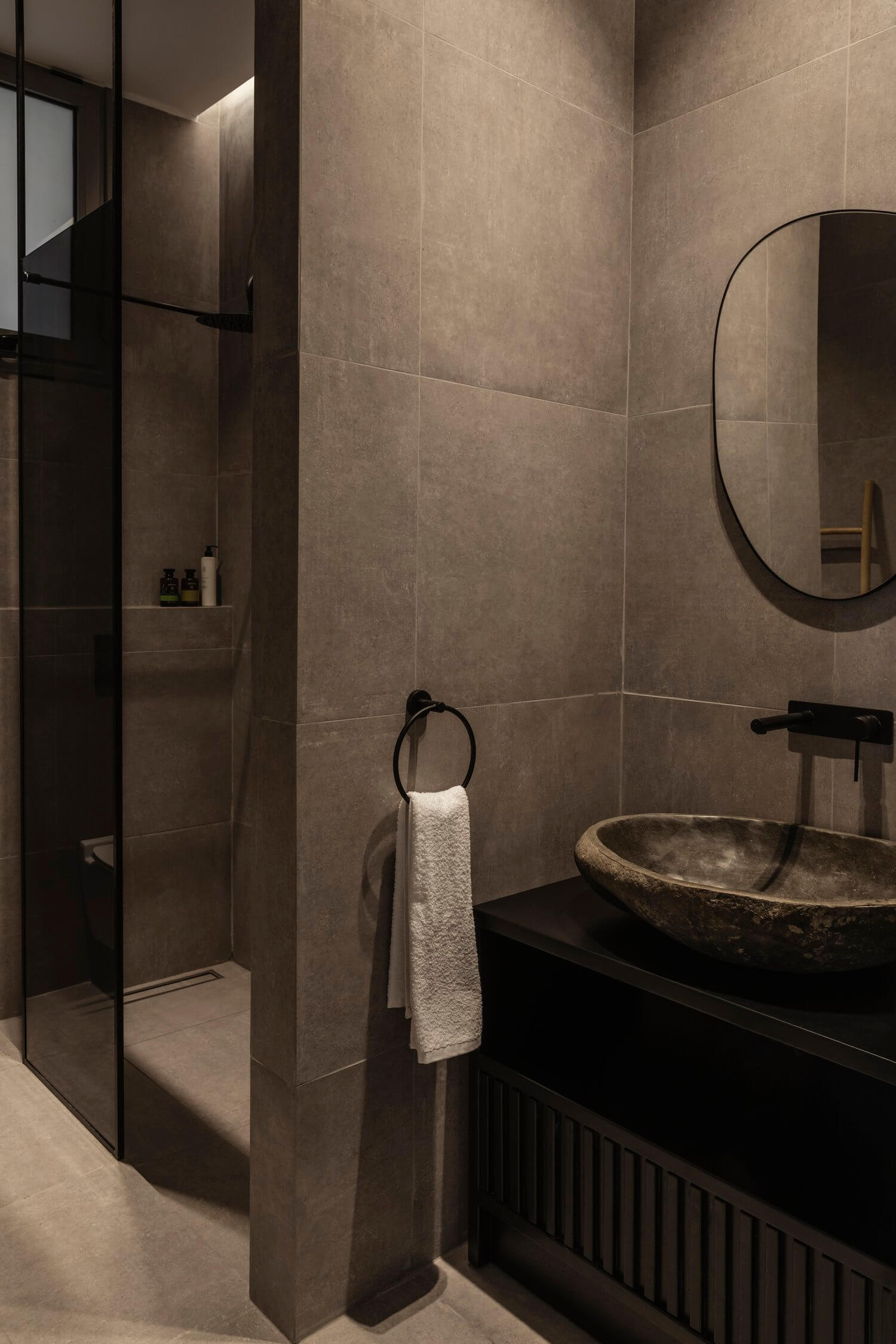 Casa Somos Suenos, Europe — Minimalist bathroom with warm taupe tile walls, black framed mirror, stone vessel sink, walk-in shower with glass partition, and natural light casting geometric shadows.