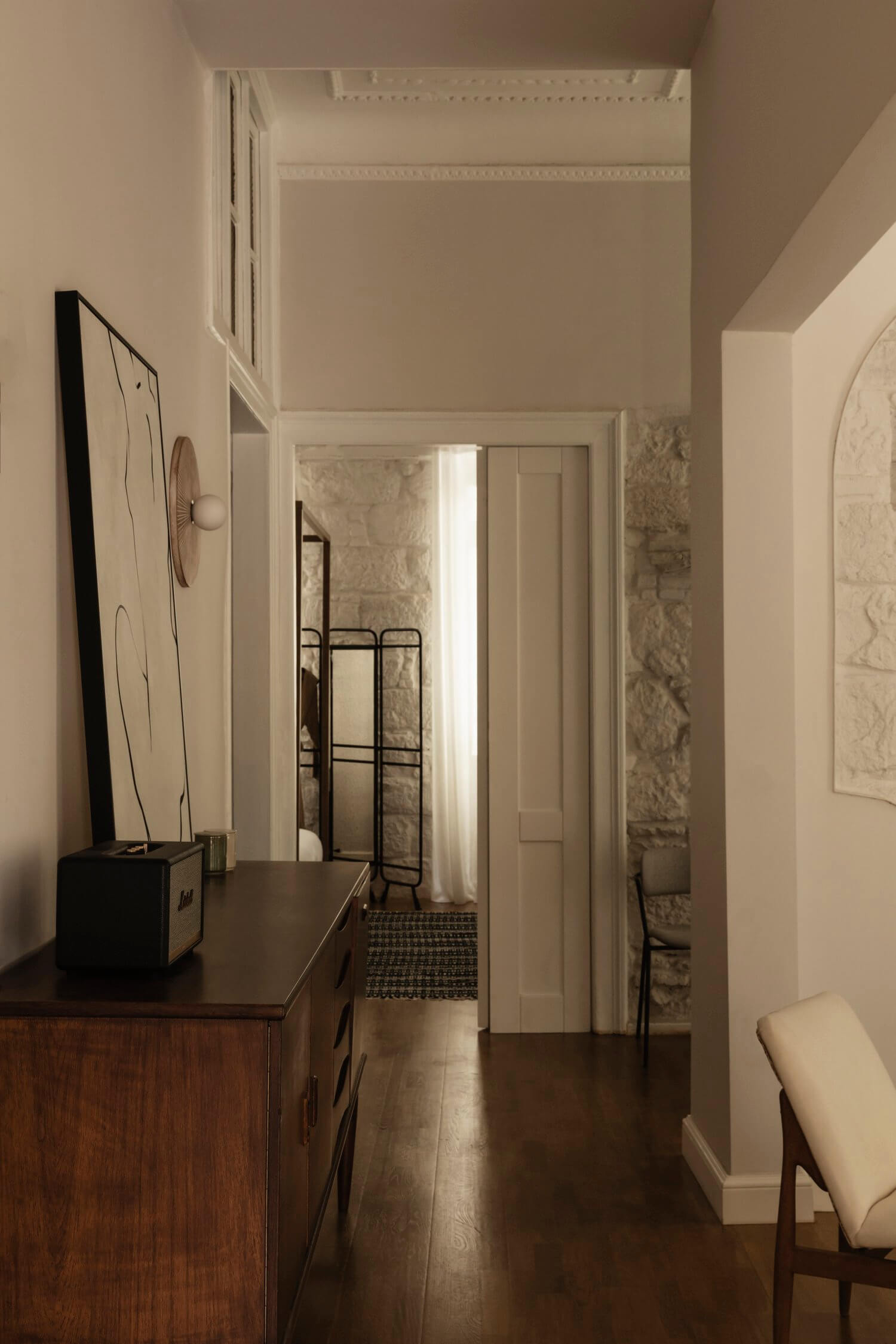 Casa Somos Sueños, Europe — Minimalist hallway with dark wood flooring, cream walls, natural light, mid-century furniture, and exposed stone archway.