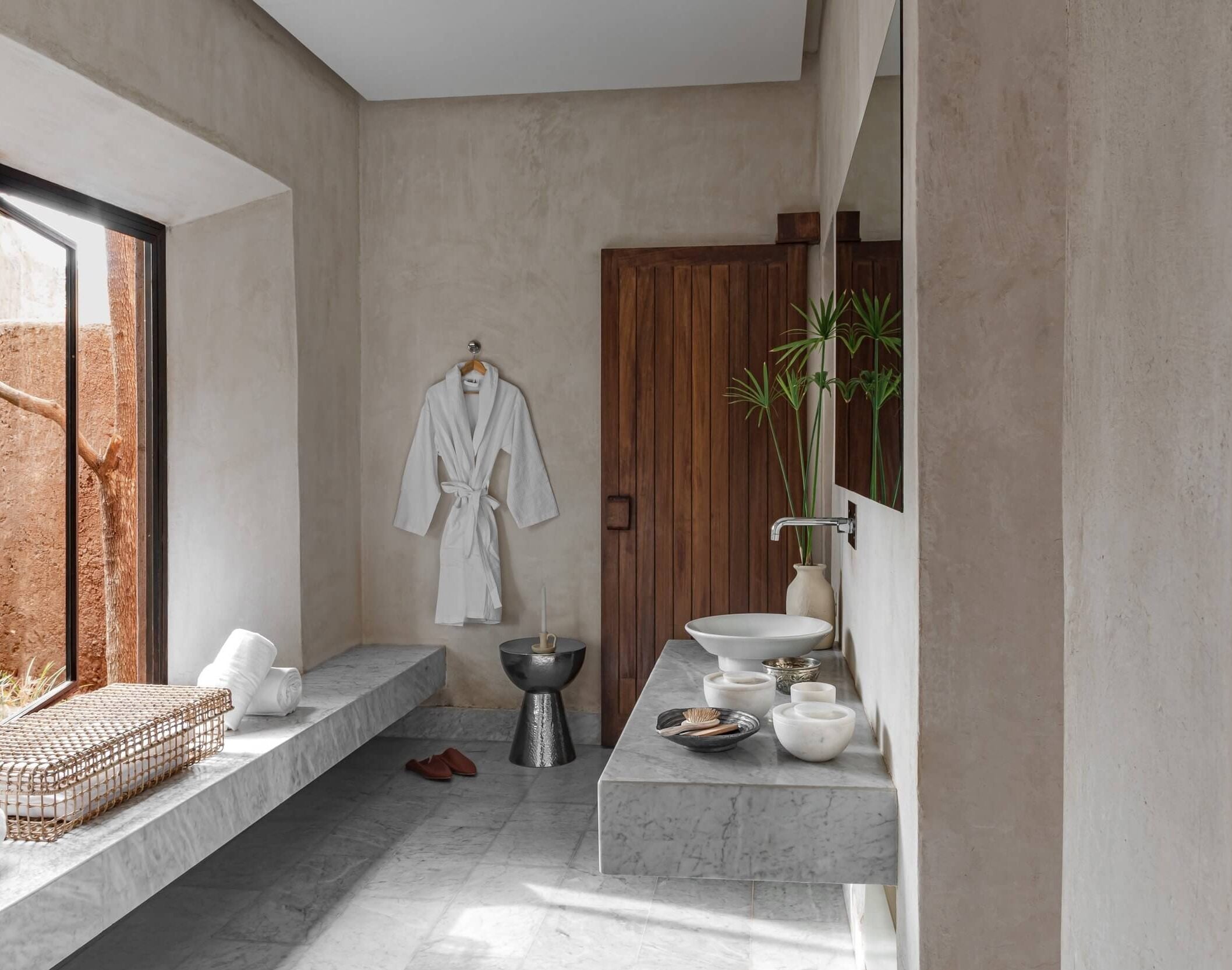 Villa D, Africa — Minimalist spa bathroom with marble counters, concrete walls, woven window seat, wooden door, vessel sink, and sculptural black accent table in natural light.