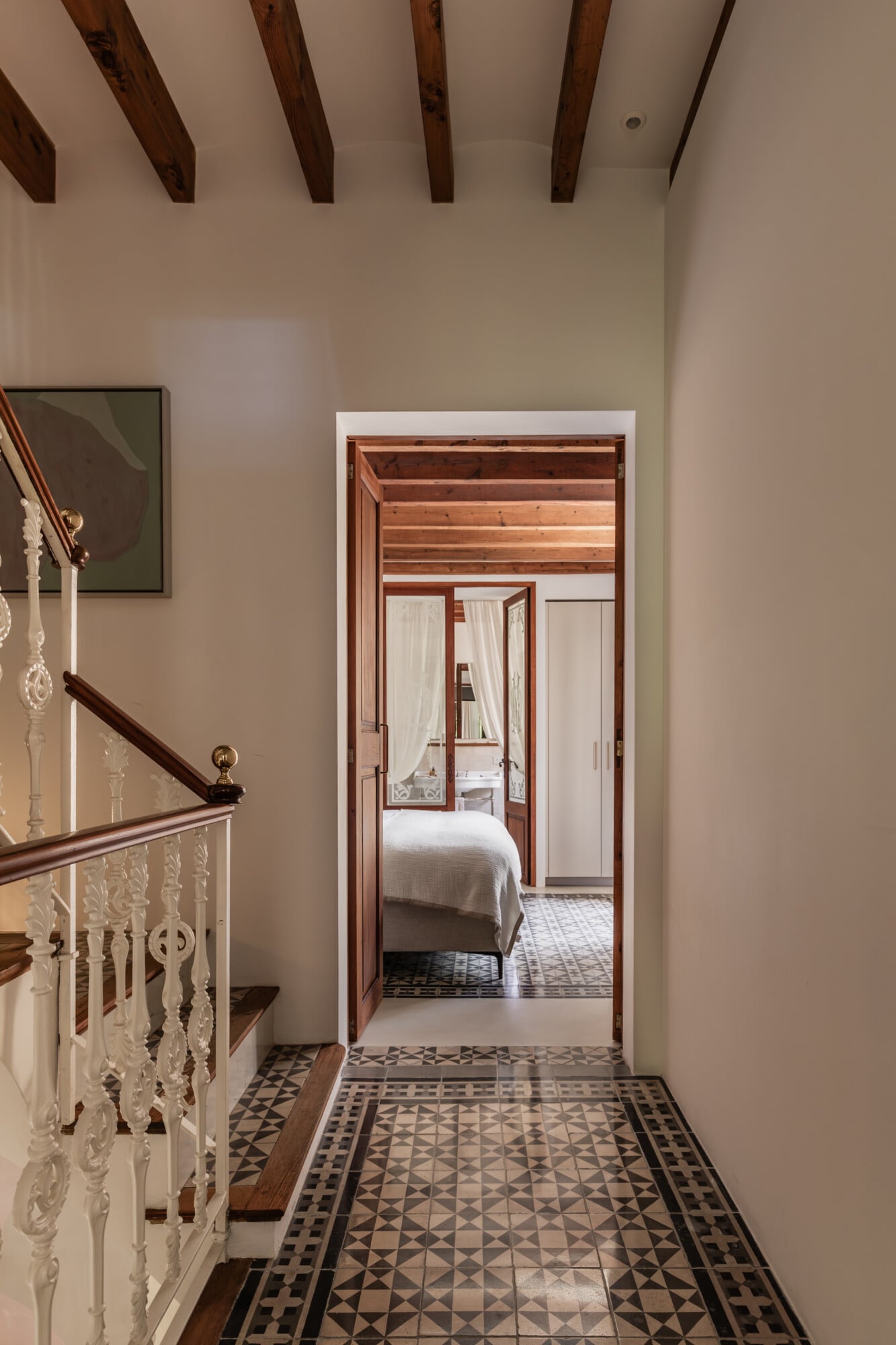 Mon Cor, Balearic Islands — Hallway with geometric tile floor, exposed wood beams, ornate railing, and open wooden doors framing bedroom beyond.