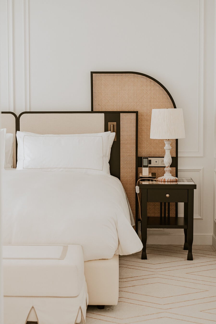 Carlton Cannes, Cannes, France — Minimalist bedroom with cream linens, black metal bed frame, rattan headboard panel, dark nightstand, and brass table lamp.