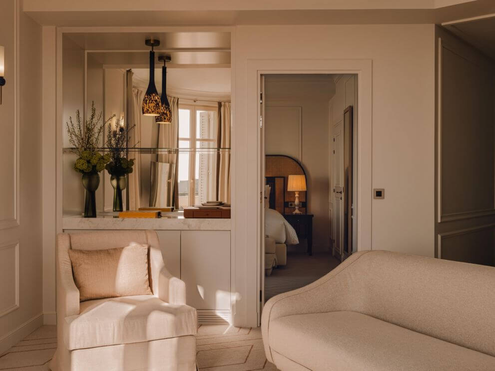 Carlton Cannes, Côte d'Azur, France — Neutral bedroom with cream upholstered seating, marble vanity, pendant lighting, and mirrored alcove overlooking Mediterranean views.