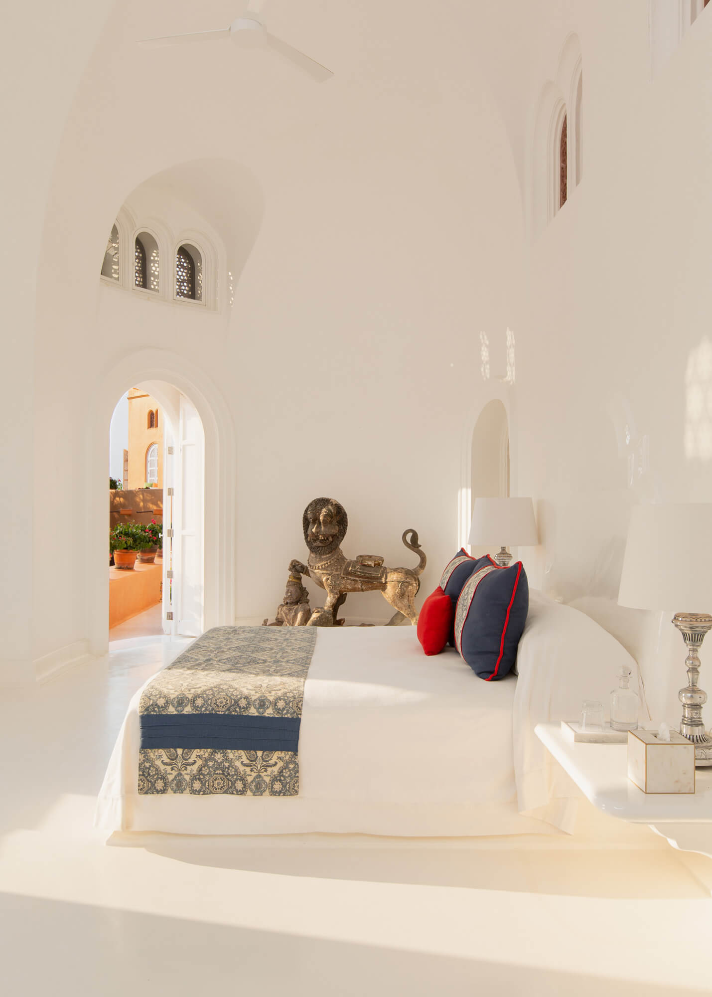 Cuixmala, Mexico — Whitewashed bedroom with arched doorways, bronze sculptural bust, patterned runner, and terracotta courtyard visible through open door.