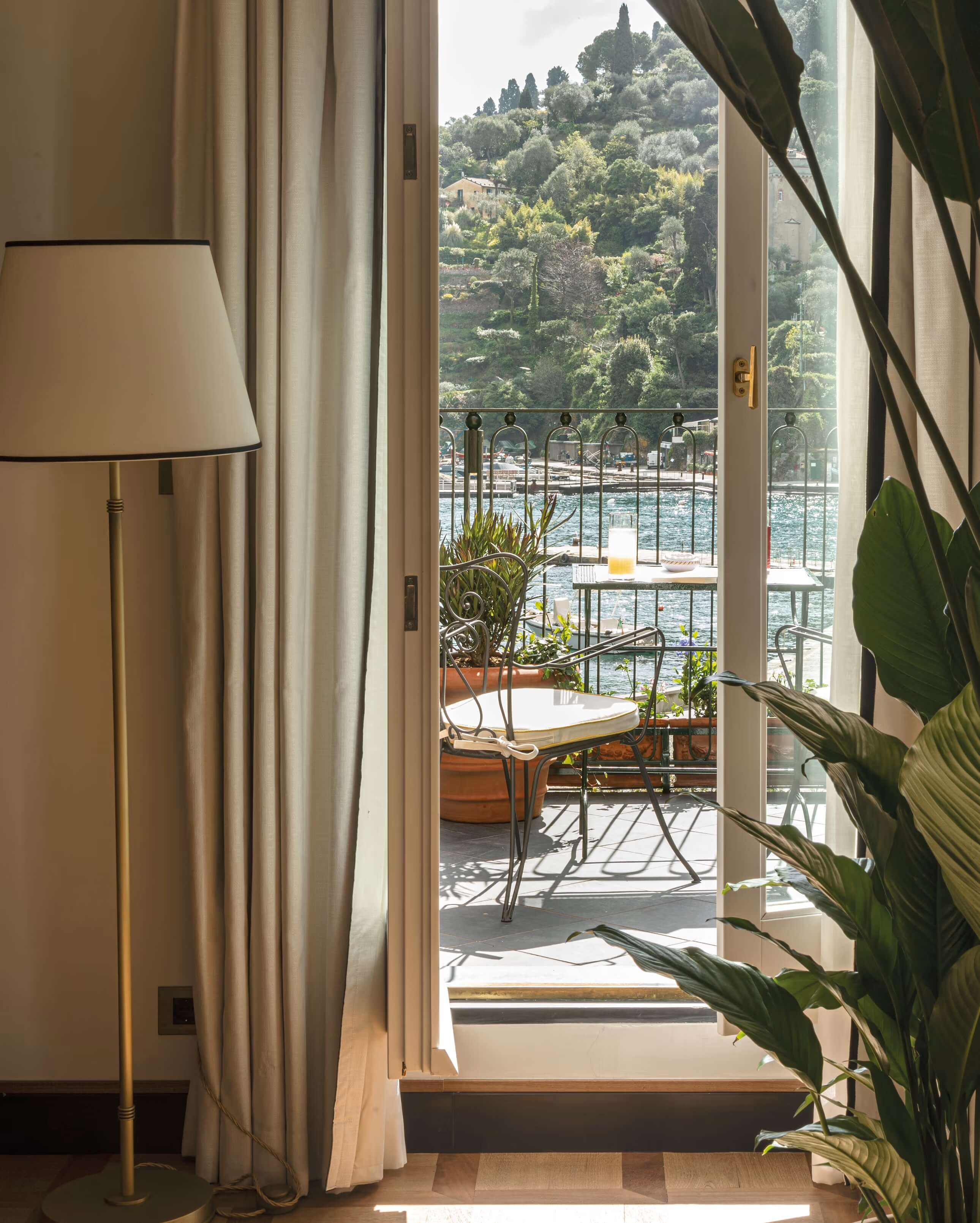 Splendido Mare, Europe — Lakeside balcony with iron railings, linen curtains, potted plants, and forested hillside views through open glass doors.