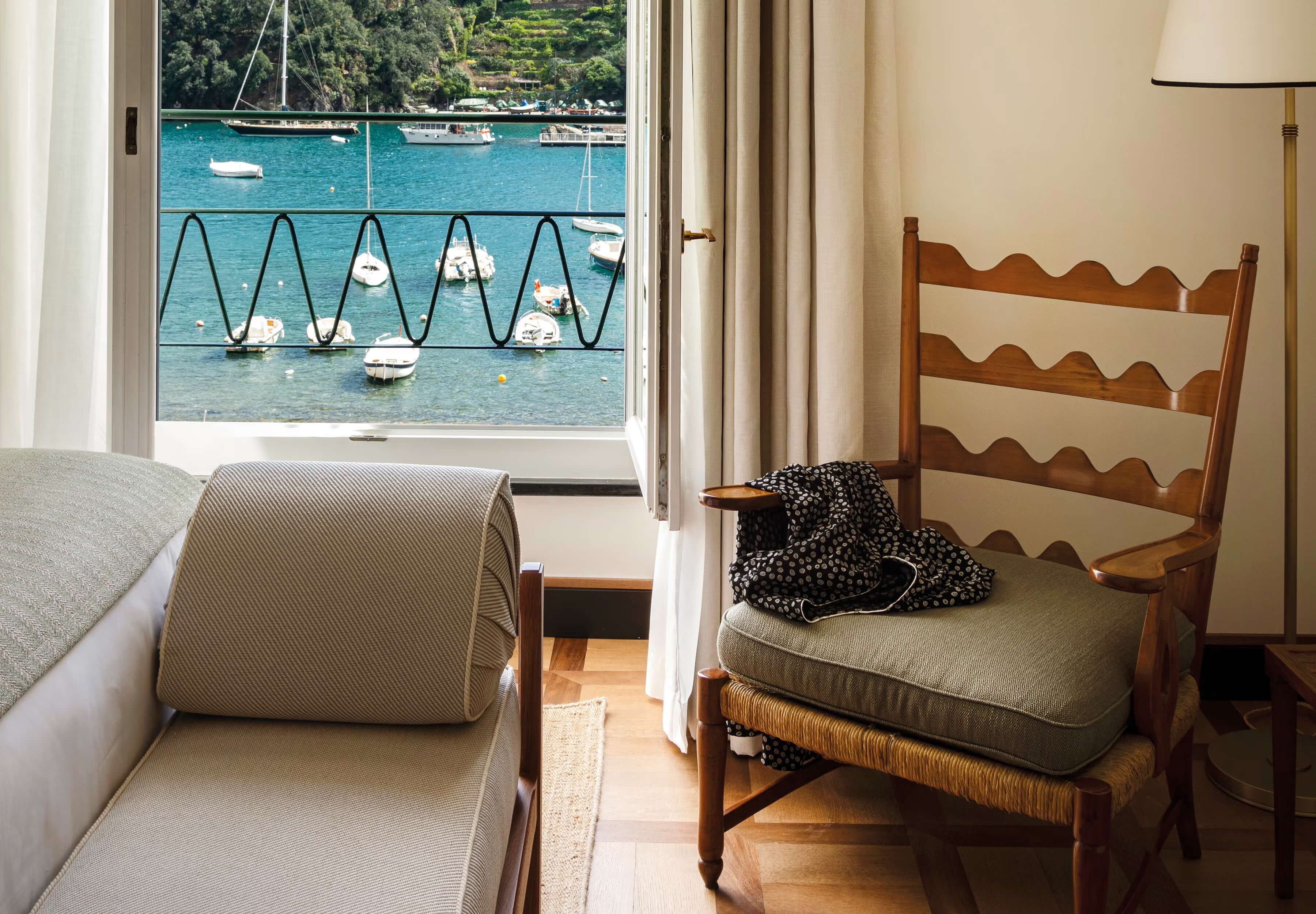 Splendido Mare, Europe — Coastal bedroom with wooden ladder-back chair, striped linen upholstery, turquoise harbor view through floor-to-ceiling windows, moored sailboats, Mediterranean landscape.