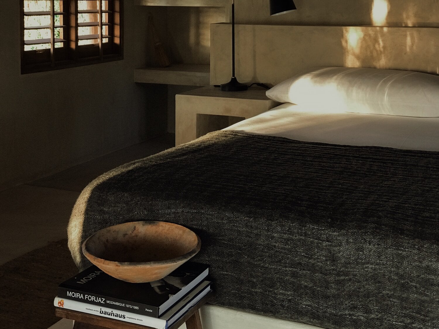Sussurro, Africa — Minimalist bedroom with black linen bed, white pillows, wooden bowl on design books, natural light through shuttered window, earth-tone walls.