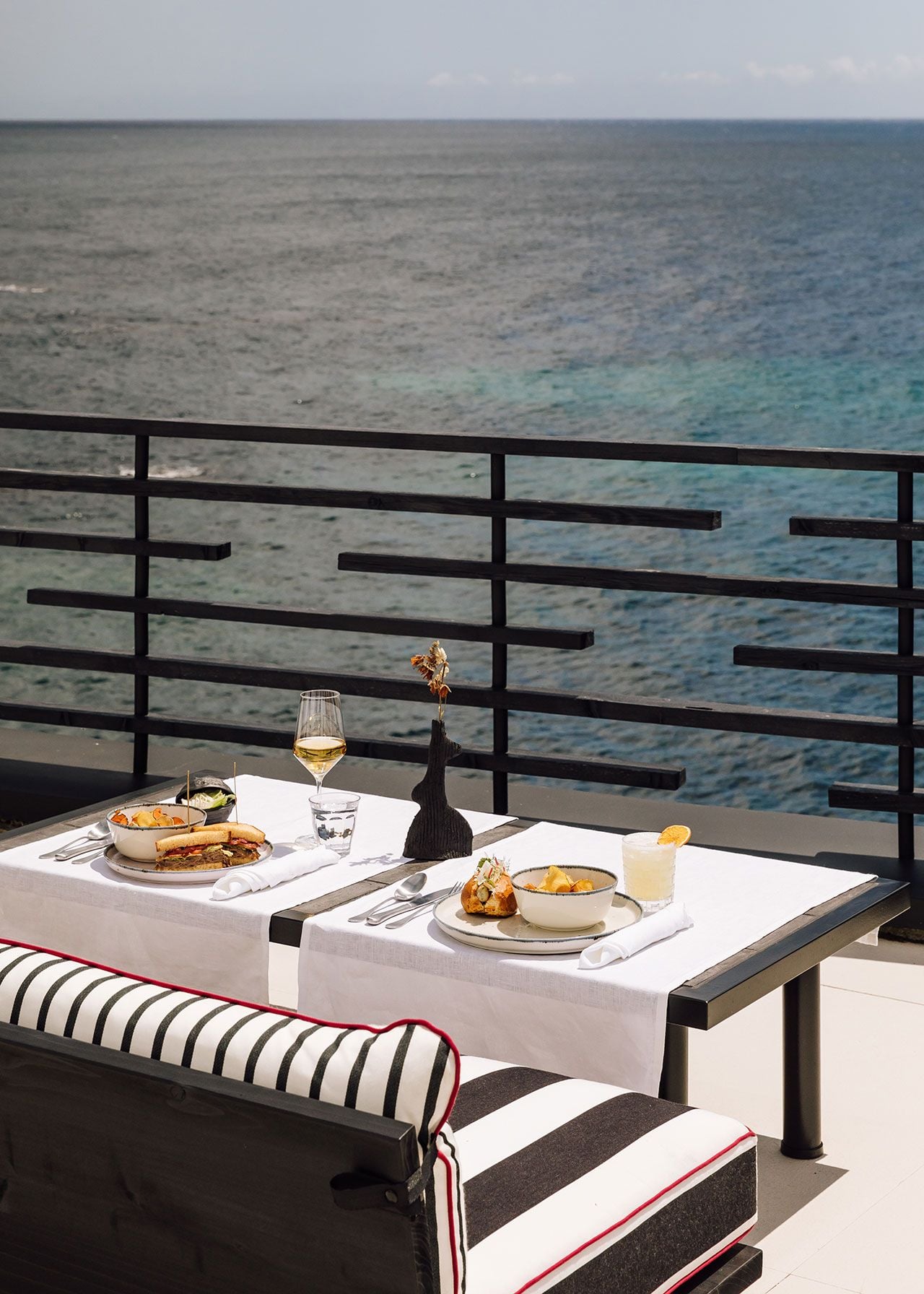 White Bar, Europe — oceanfront dining table set with white linens, wine glass, and plated cuisine overlooking turquoise waters beyond black geometric railing.